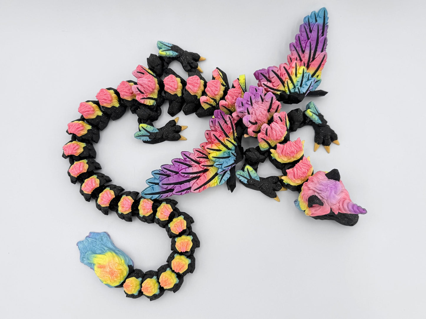Top view of a 3D printed Unicorn Dragon Fidget in glitter black with rainbow wings and mane. The articulated body is coiled into a spiral shape, showing flexible joints and detailed layered scales.