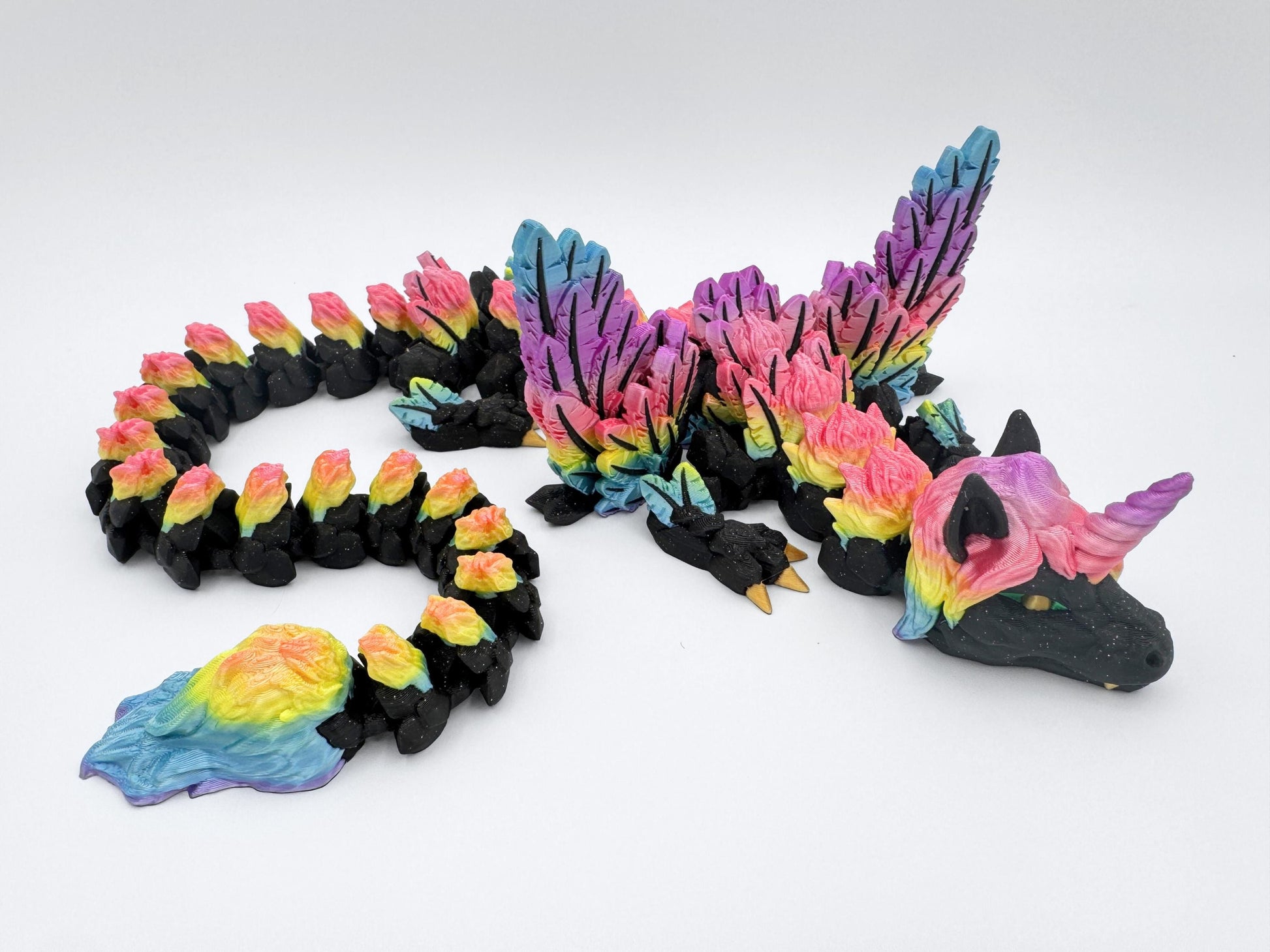 Unicorn Dragon articulated figurine 3D printed in rainbow and glitter black. The dragon is posed with its body curled, highlighting colorful tail segments, feather wings and a rainbow unicorn horn.