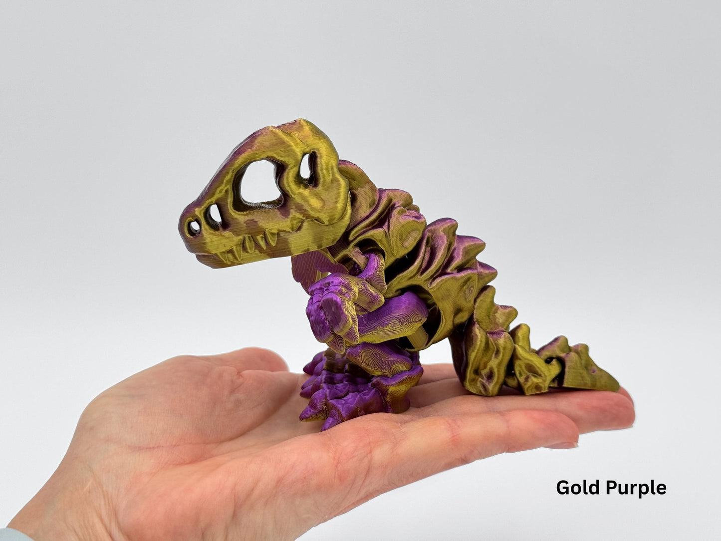 Gold and purple articulated dinosaur fidget figurine displayed on a hand. The 3D printed T-rex style dragon has a large head, jointed limbs and a curled segmented tail with smooth movement.