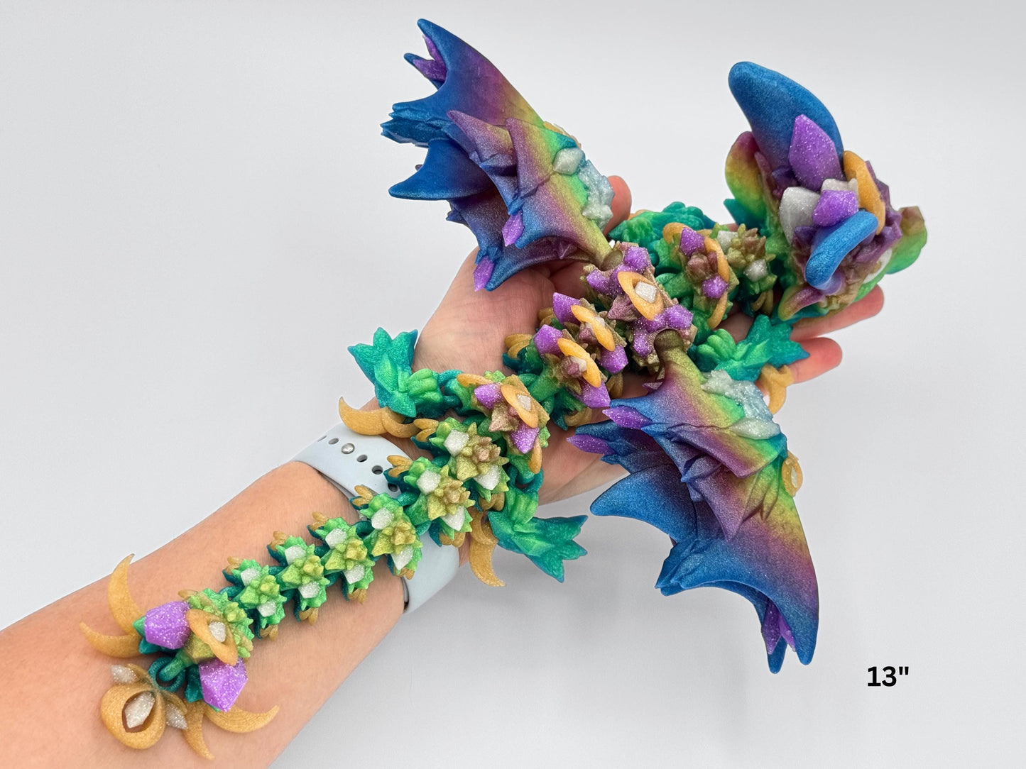 3d printed night wing dragon figurine resting in hand for scale. Printed in rainbow colors with purple spines, teal wings, and gold ringed flowers, this 13” articulated dragon fidget has a fantasy creature design and flexes along its length. Crystal details are visible along the back.