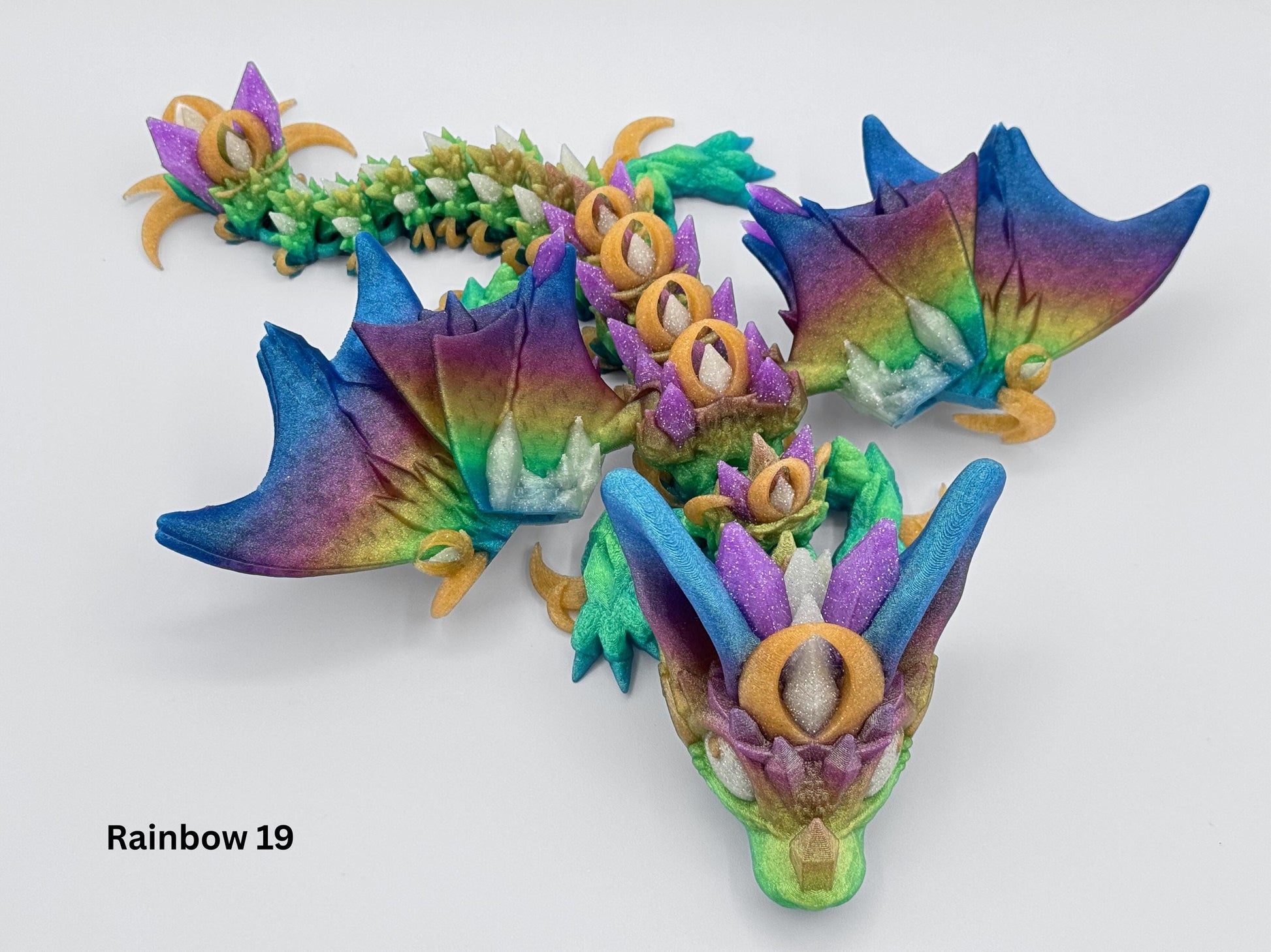 Full top down view of a 3D printed night wing dragon figurine in a flexible pose. This articulated dragon fidget features layered wings, crystal accents and a spectrum of jewel toned colors including blue, gold, green and violet. Dragon is printed in a fantasy style with flowing motion along its body, suitable as a display collectible or fidget desk piece.
