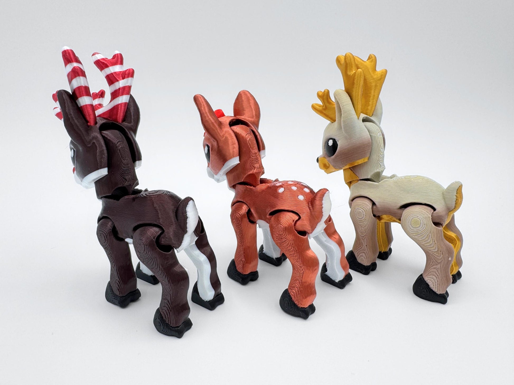Back view of three 3D printed reindeer figurines with articulated joints. The tan reindeer has golden antlers, the copper red reindeer has a red bow and white spots on its back, and the dark brown reindeer has red and white candy cane antlers. These articulated reindeer can be used as Christmas display figurines, stocking stuffers, or fidget collectibles.