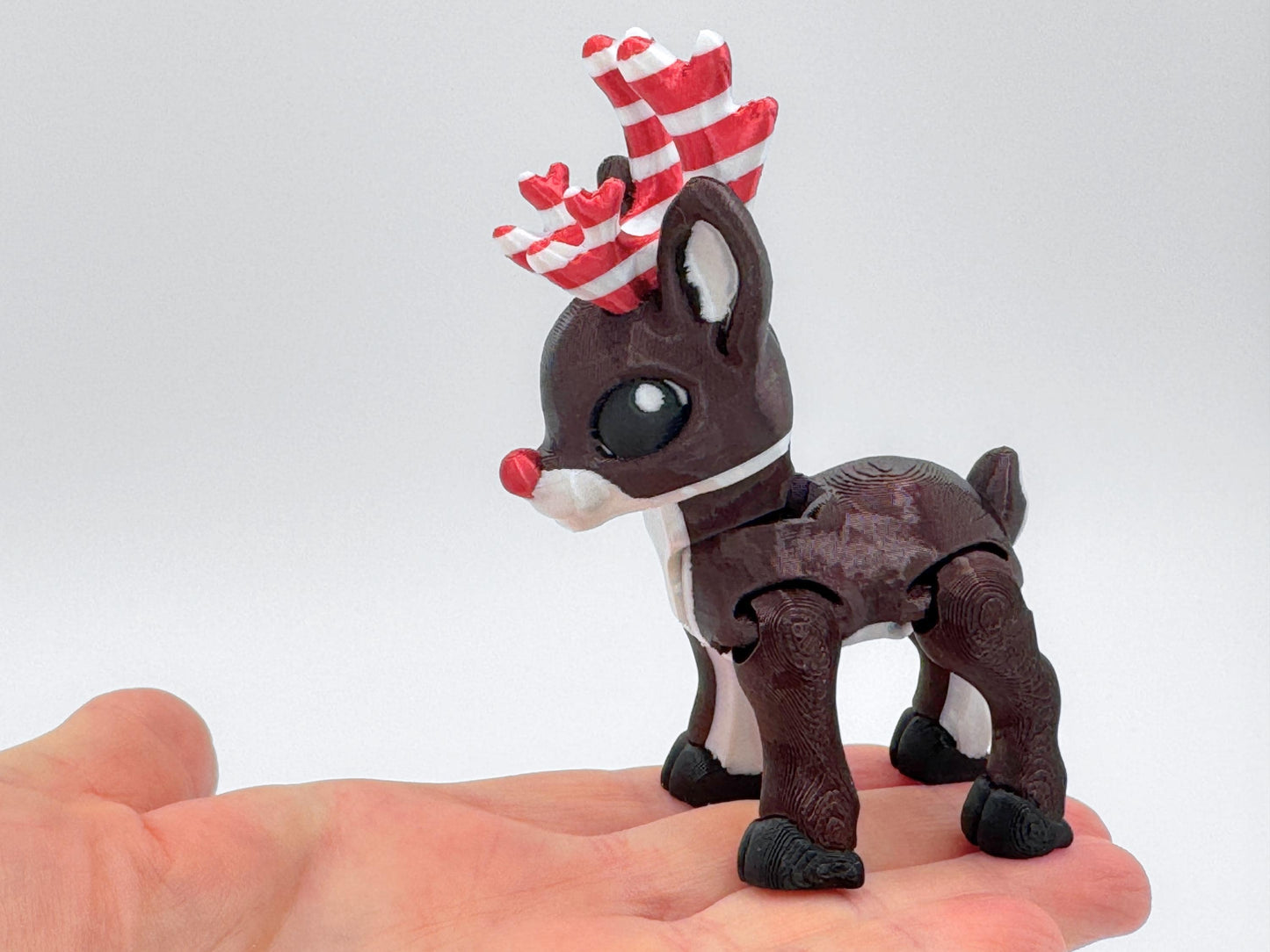 Close-up of a dark brown articulated reindeer figurine with candy cane red and white striped antlers, posed in a hand to show size. The reindeer has a red nose, black hooves, and big black eyes, with a shiny silky finish. This 3D printed reindeer can be used as a Christmas decor or festive collectible.
