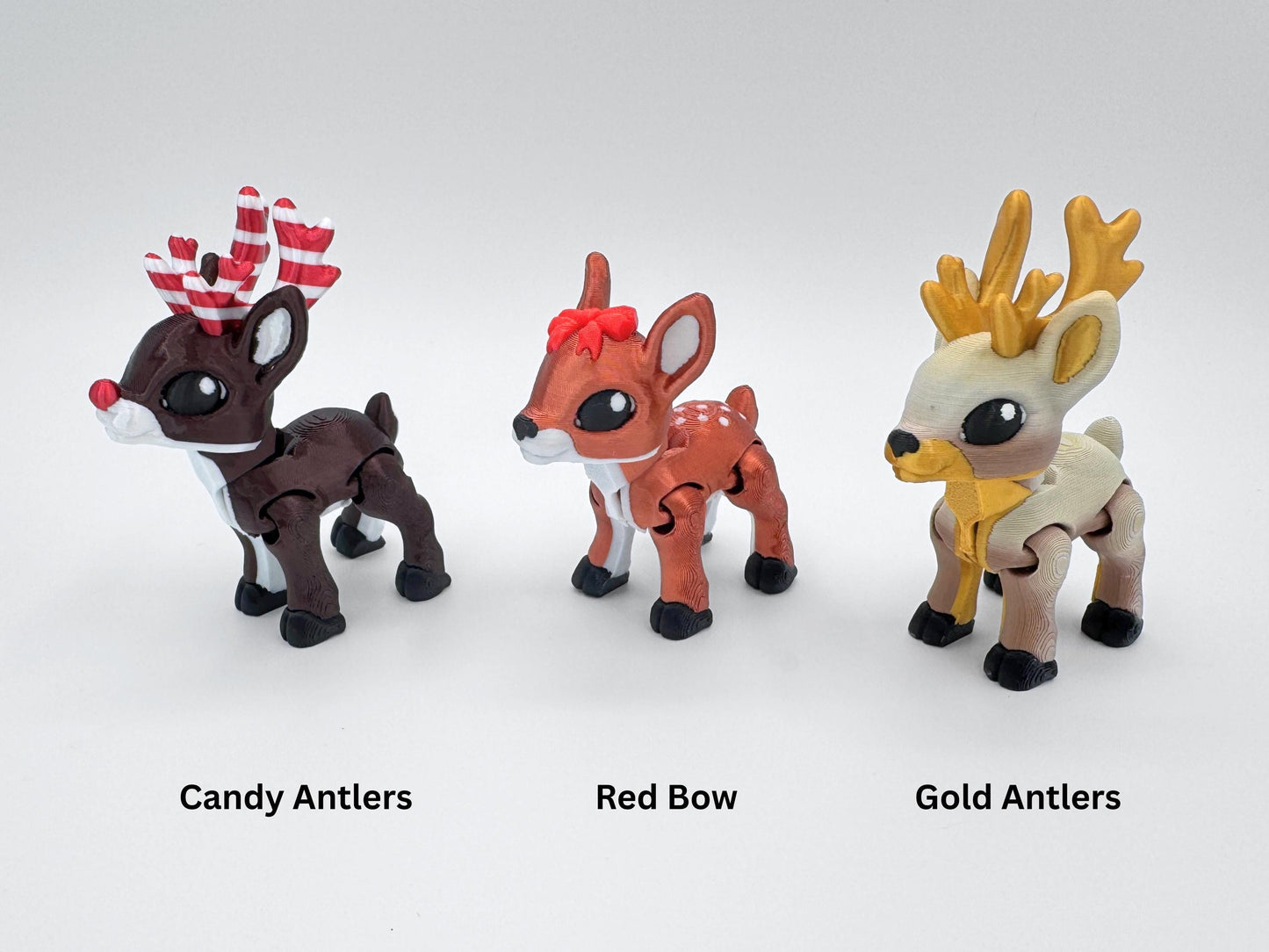 Front view of three articulated reindeer figurines standing in a row. The tan reindeer has golden antlers, the copper red one has a red bow and the dark brown reindeer has red and white striped antlers. Each reindeer has black hooves and expressive eyes, shown as Christmas fidget figurines or festive holiday collectibles.