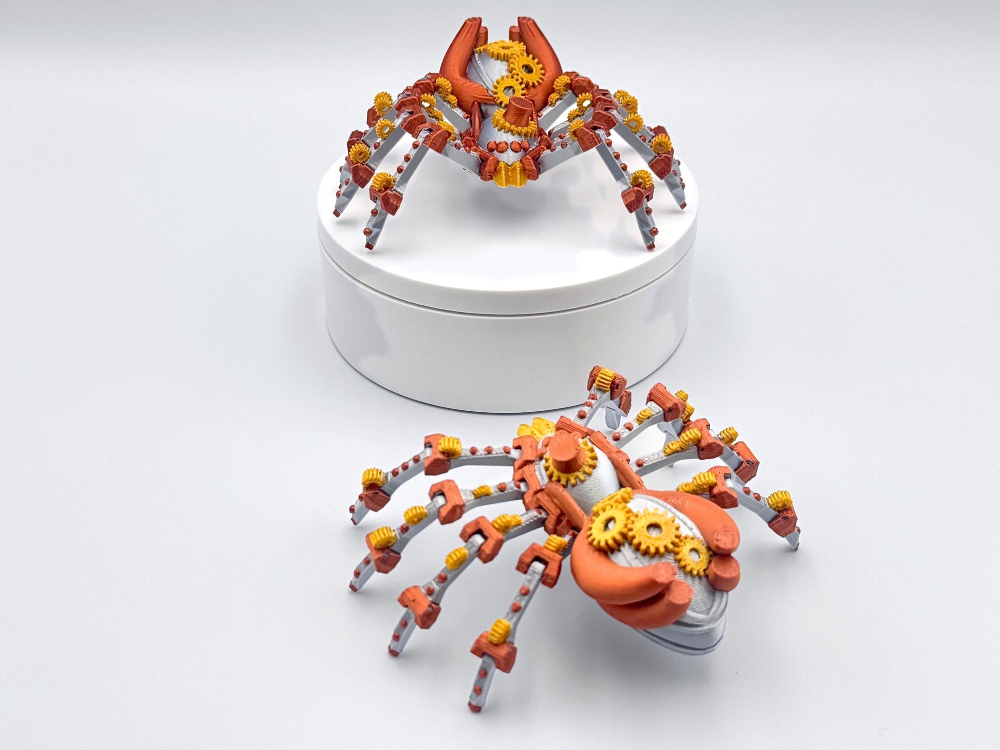 Two 3D printed, steampunk spider figurines in silver, copper and gold colors. The spiders have intricate mechanical details, including gears and are posed on a white surface.