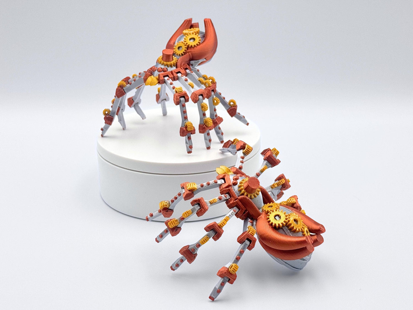 Two silver and copper 3d printed mechanical steampunk spiders with gold gears. The spiders have eight legs and are posed on a white surface.