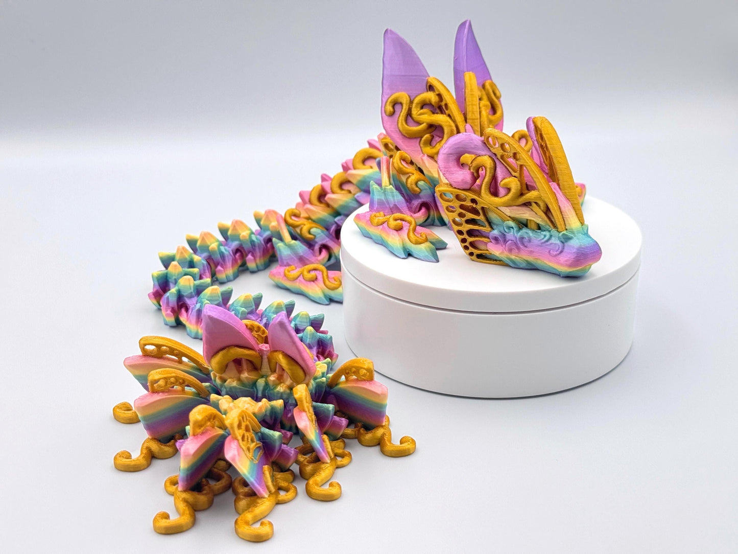 3D printed butterfly dragon figurine in rainbow filament with gold accents and details, shown curled on a white stand with articulated segments and sculpted texture.