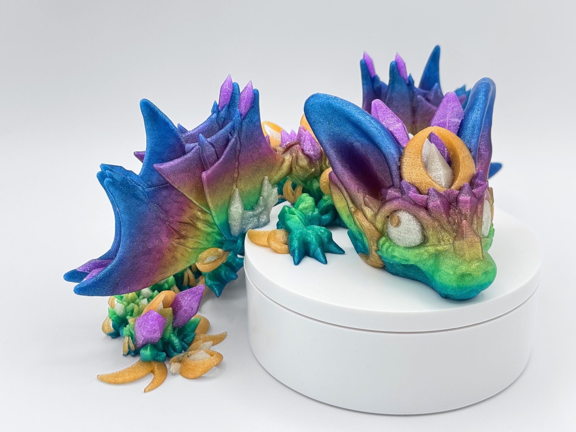 Colorful articulated night wing dragon figurine shown curled around a white circular base. Features bright rainbow gradient with vivid purple, teal, gold and green tones. The dragon has horn like ears and crystal along its back, creating a fantasy inspired display. 3D printed in multiple filament colors with detailed textures and flexible joints for fidgeting or posing.