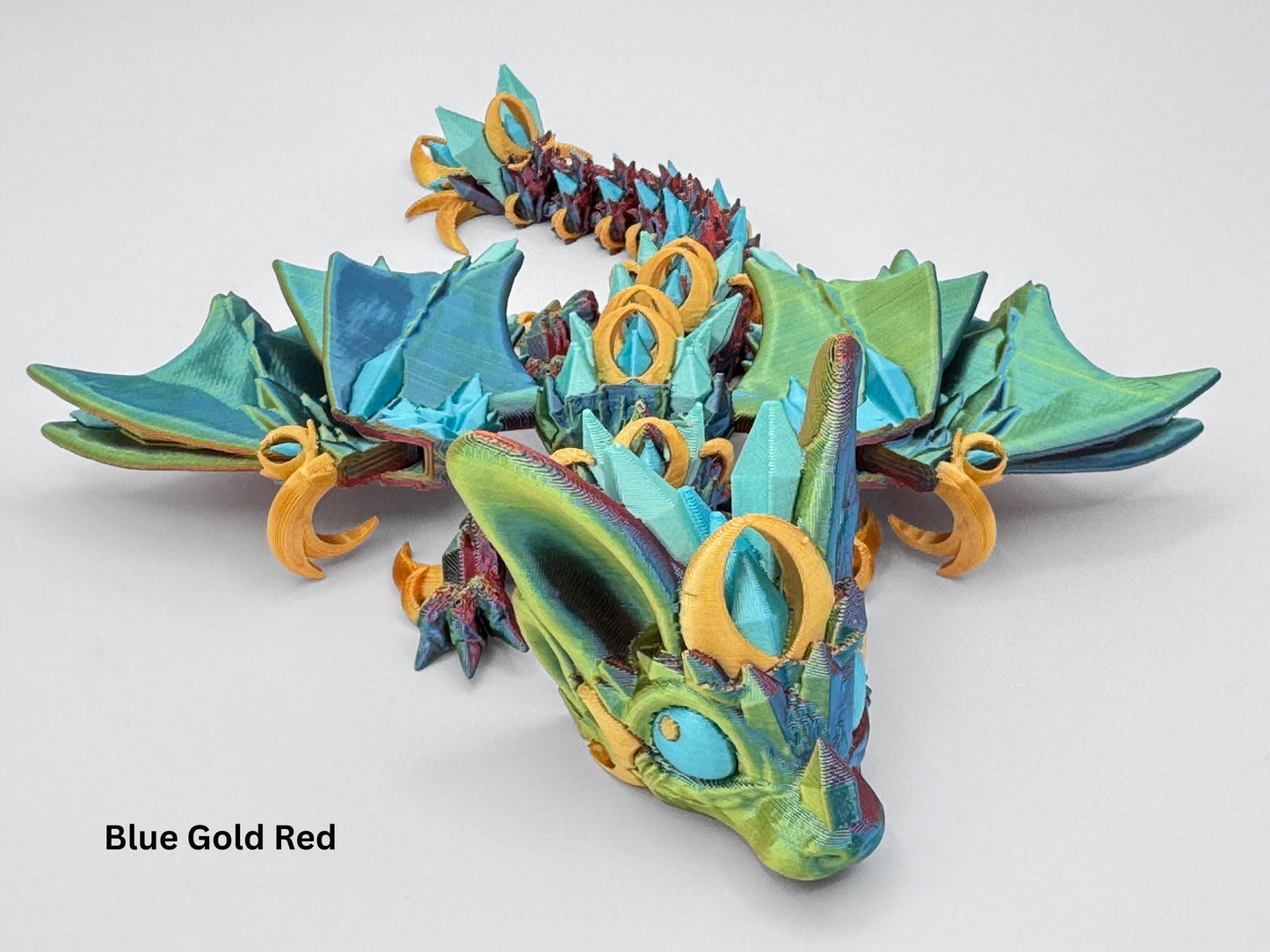 Night Wing Dragon in tri-color blend, displayed on a tabletop. Dragon has flexible, poseable body with teal and blue crystal-like ridges along its back, ear-shaped side details, and sweeping articulated wings. Color gradient on its body transitions from deep red to blue and yellow. Shown curled in a relaxed pose to highlight movement and detailed texturing.