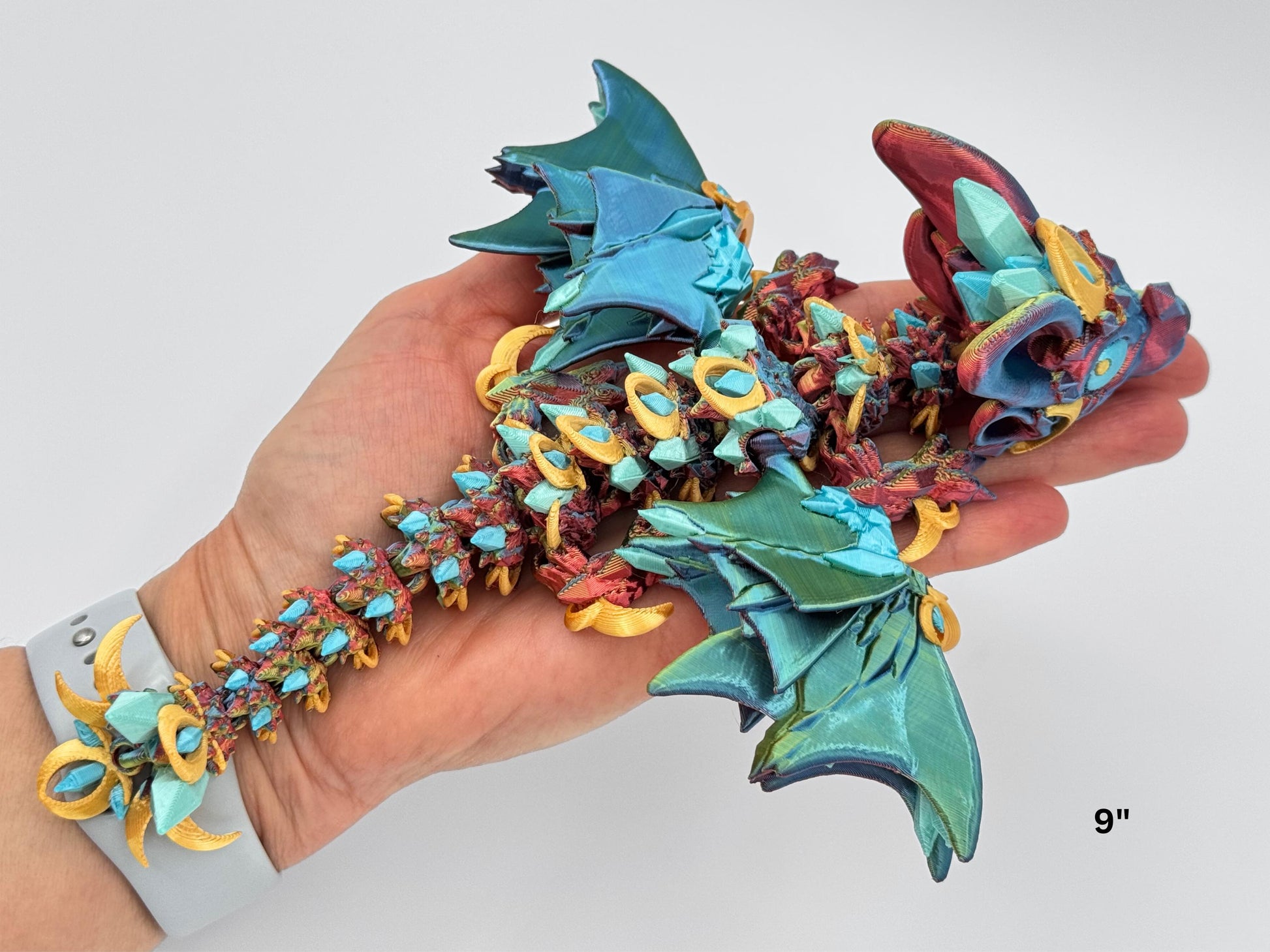 9” Night Wing Dragon held in hand to show size. This articulated 3D printed figurine features moon-shaped gold accents, ear-shaped side details, and a crystal-textured back in teal and blue. The tri-color body fades from red to blue to yellow, with flexible joints and interlocking rings that enhance its fidget-style movement.