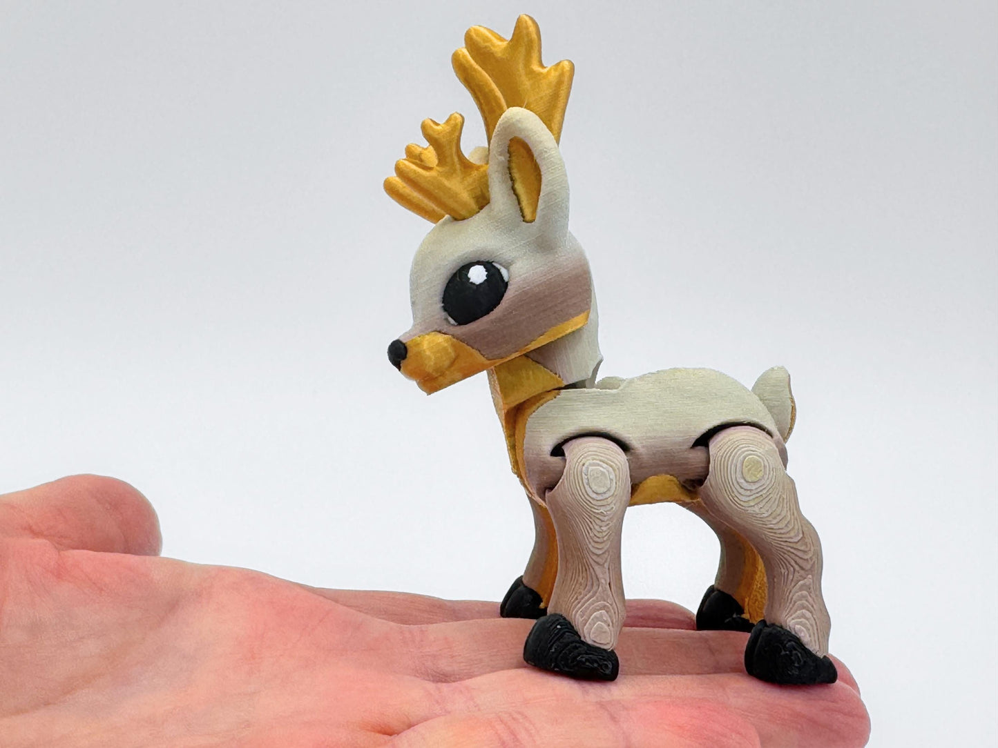 Close-up of a tan 3D printed reindeer figurine with golden antlers, resting on a hand to show size. The reindeer has articulated legs, black hooves, and eyes. This reindeer works as a small holiday decoration, Christmas collectible, or articulated fidget figurine.
