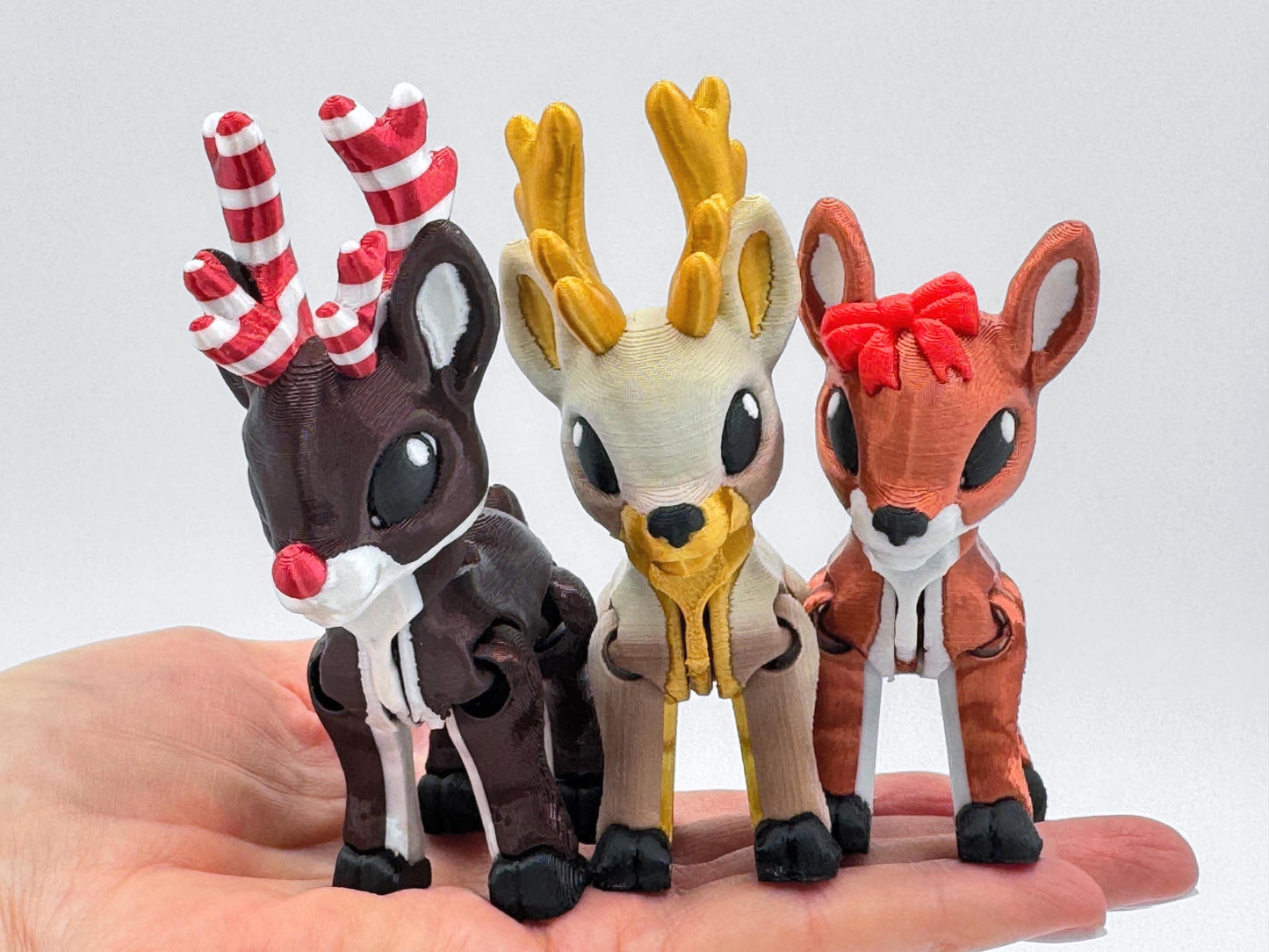 Three holiday reindeer figurines held together in one hand, showing the variety of designs. One has golden antlers, another has candy cane striped antlers, and the third has a red bow. Each articulated reindeer has flexible legs and black hooves, shown as Christmas collectibles and festive holiday display decor.