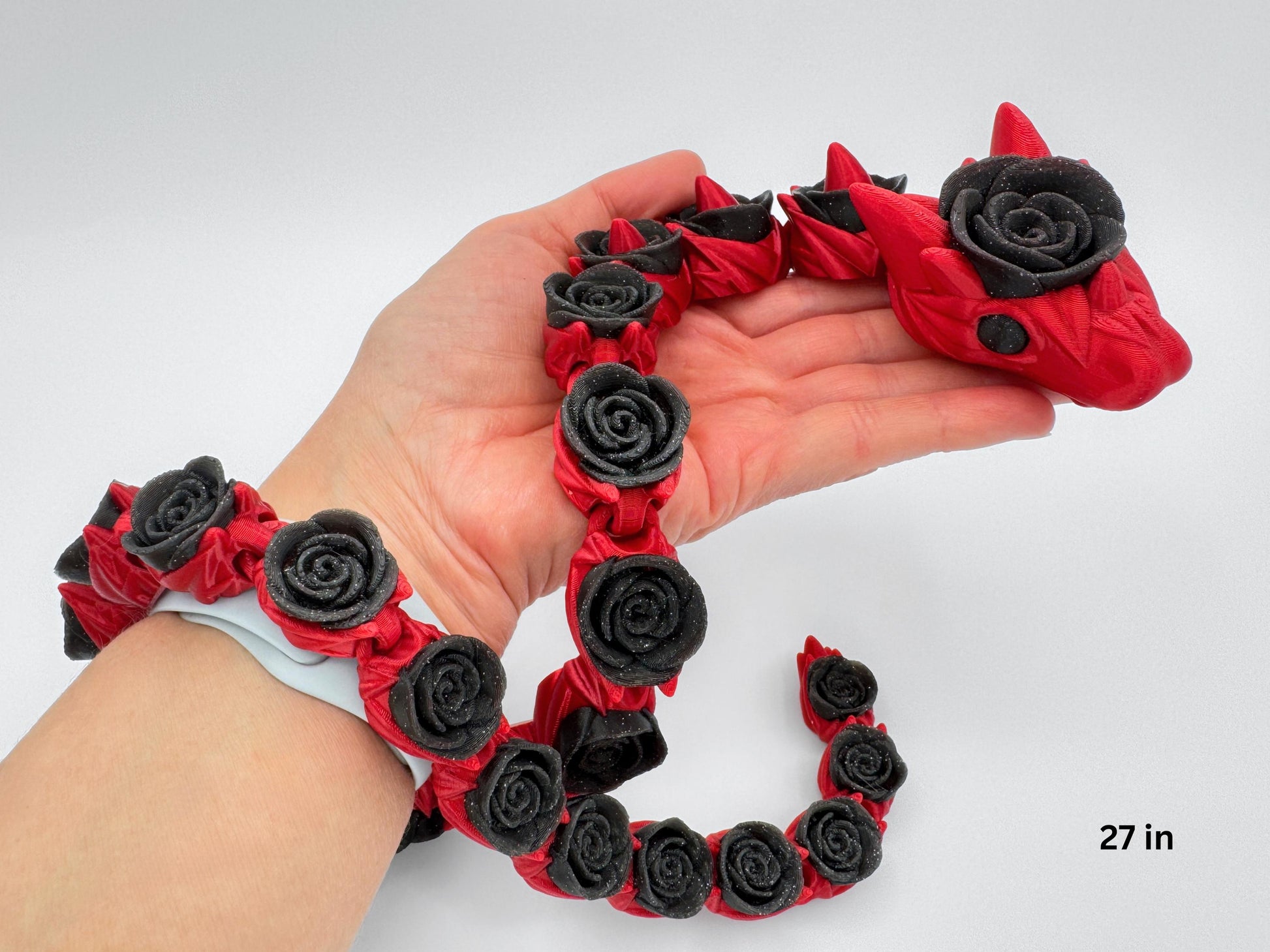 Articulated rose snake held in a hand for scale. The long, slender body drapes across fingers, showing how the linked segments bend easily. Red body with glitter-black rose-shaped details runs from head to tail. The in-hand view provides size context, reveals surface texture, and emphasizes the fidget’s flexible motion against a simple background.