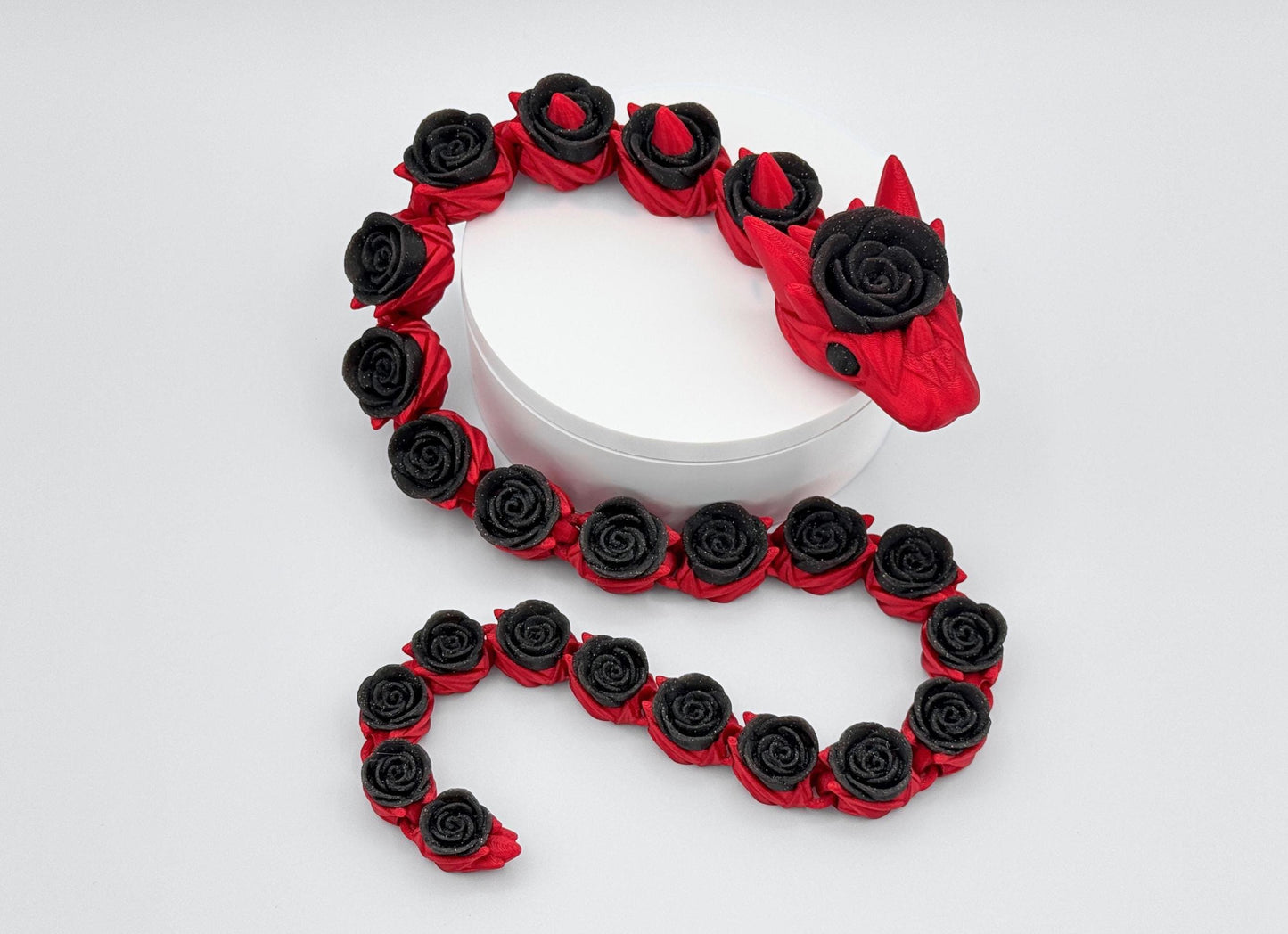 Red-bodied, glitter-black rose-segmented articulated rose snake posed in a loose coil on a white stand. The head faces forward; repeating rose shapes run the length of the flexible body. Gentle S-curve shows how the linked sections bend and settle naturally, with smooth surface detail and distinct color separation against a neutral background.