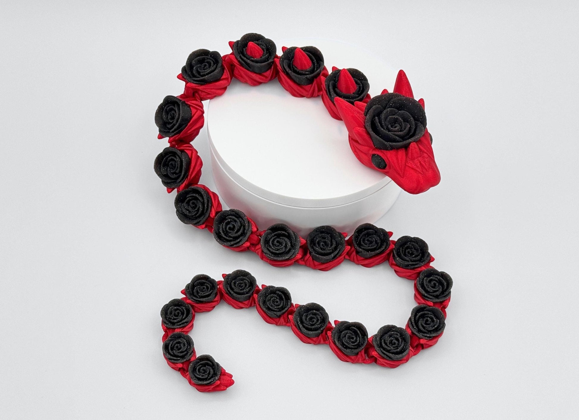 Red-bodied, glitter-black rose-segmented articulated rose snake posed in a loose coil on a white stand. The head faces forward; repeating rose shapes run the length of the flexible body. Gentle S-curve shows how the linked sections bend and settle naturally, with smooth surface detail and distinct color separation against a neutral background.