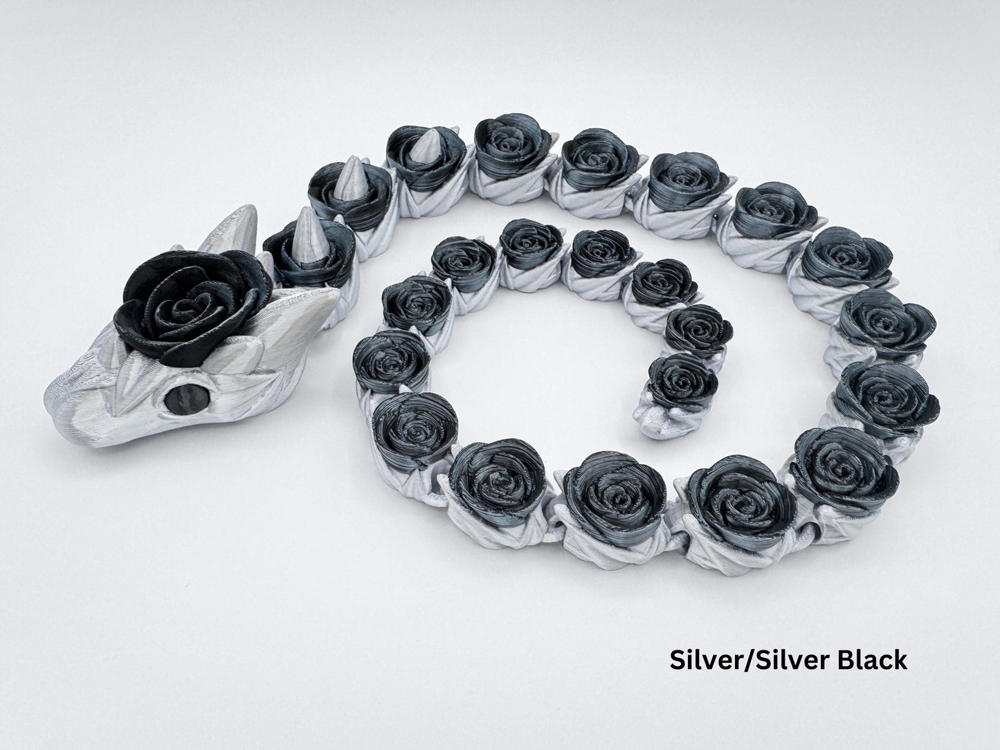 3D printed articulated rose snake fidget in silver and silver black, posed in a loose spiral on a white background. The body is silver with dark rose-shaped segments along the length and a matching rose on the head; small conical spikes are visible near the neck. On-image text reads “Silver/Silver Black.