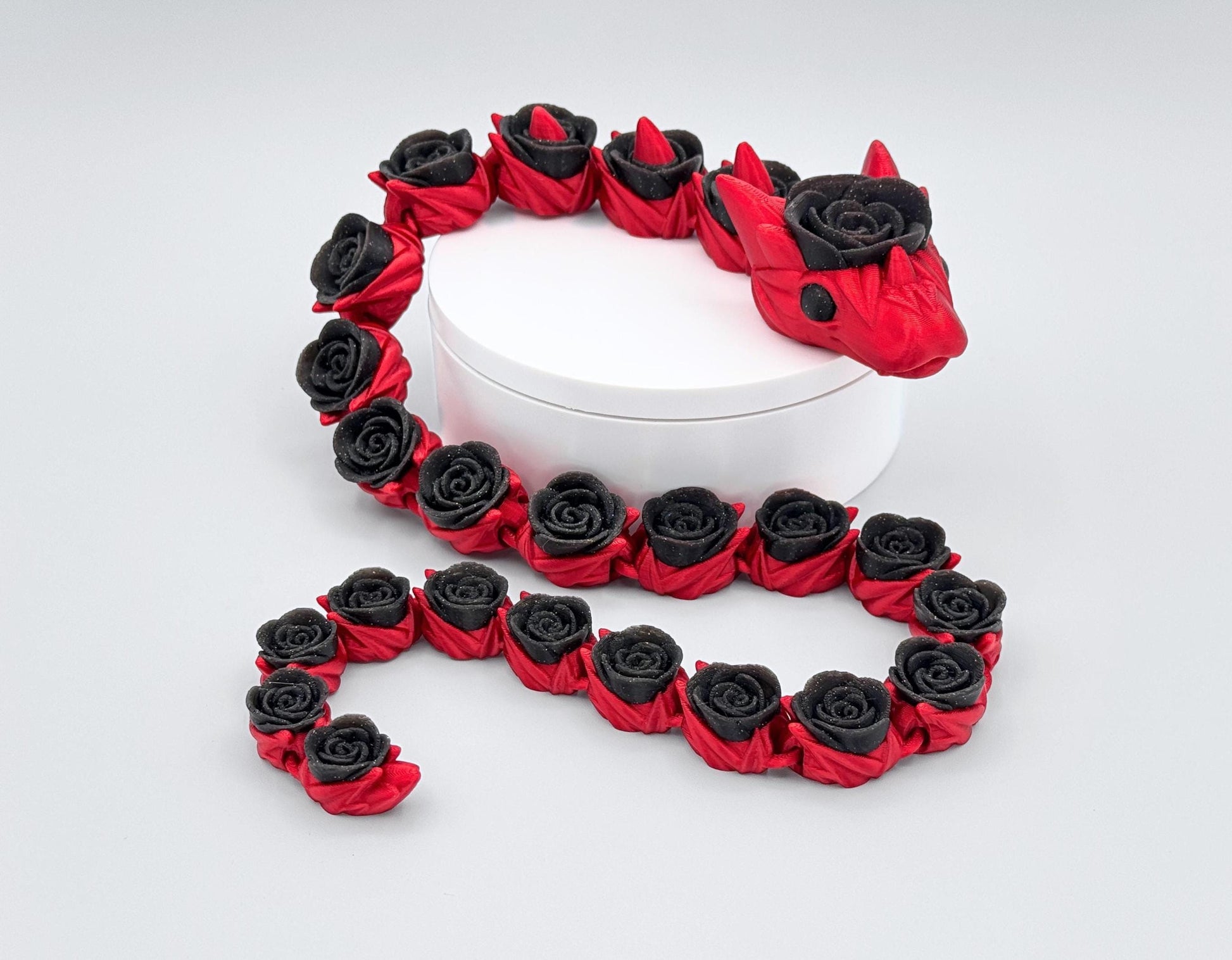 3D printed articulated rose snake fidget coiled around a white circular stand. The body is red with glitter-black rose-shaped segments linked along the spine. Curved pose emphasizes the flexible joints, smooth surface texture, and high-contrast color pattern. Neutral white background keeps focus on the snake and the sculpted rose details.