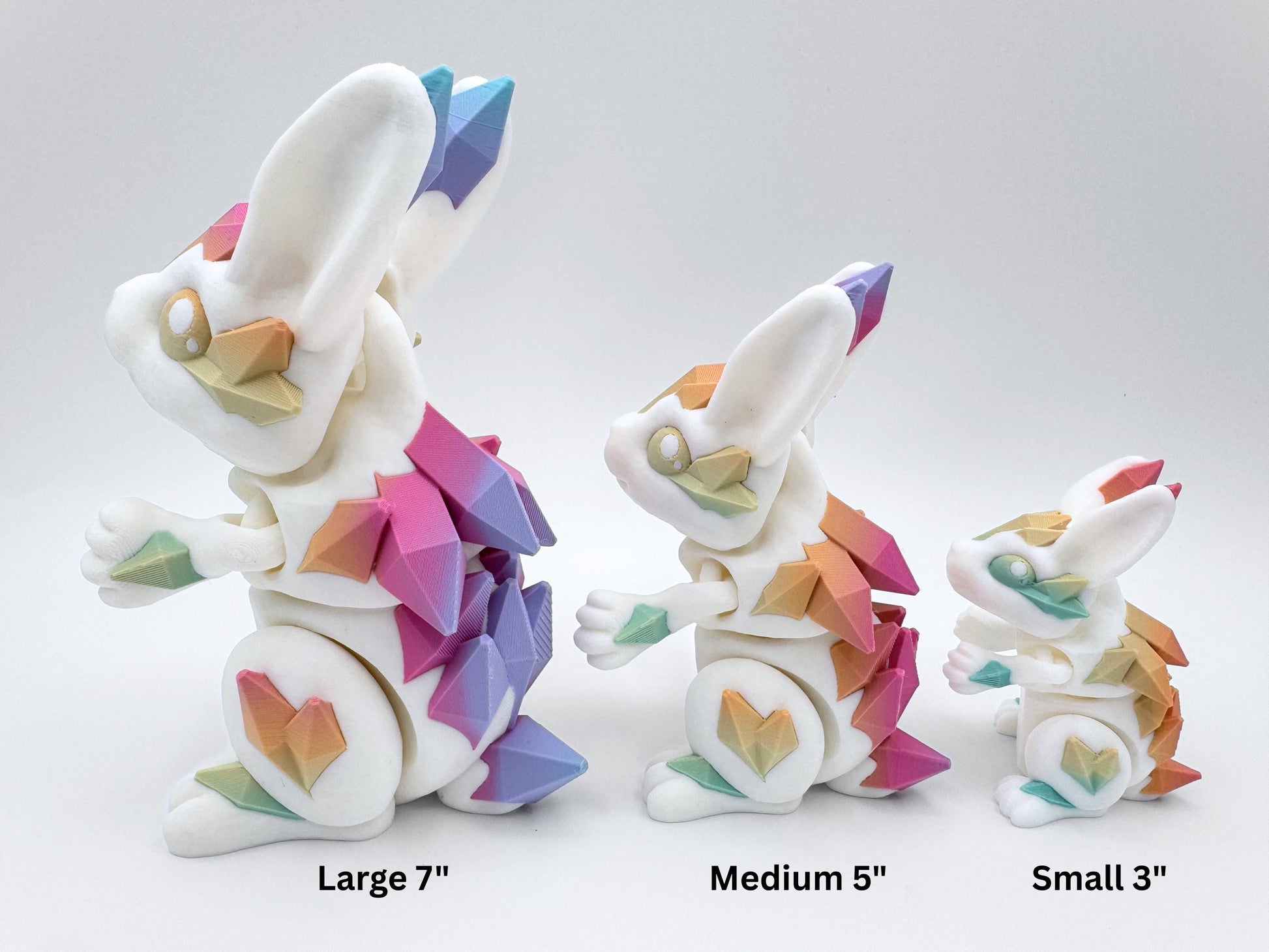 Side-by-side size comparison of three articulated Crystal Bunny figures, 3D printed in white with matte pastel rainbow crystal plates. From left to right the bunnies are Large, Medium, and Small, shown in profile against a plain background. The crystals gradate pink, purple, blue, teal, yellow, and peach. Overlay labels beneath each read “Large 7",” “Medium 5",” and “Small 3".