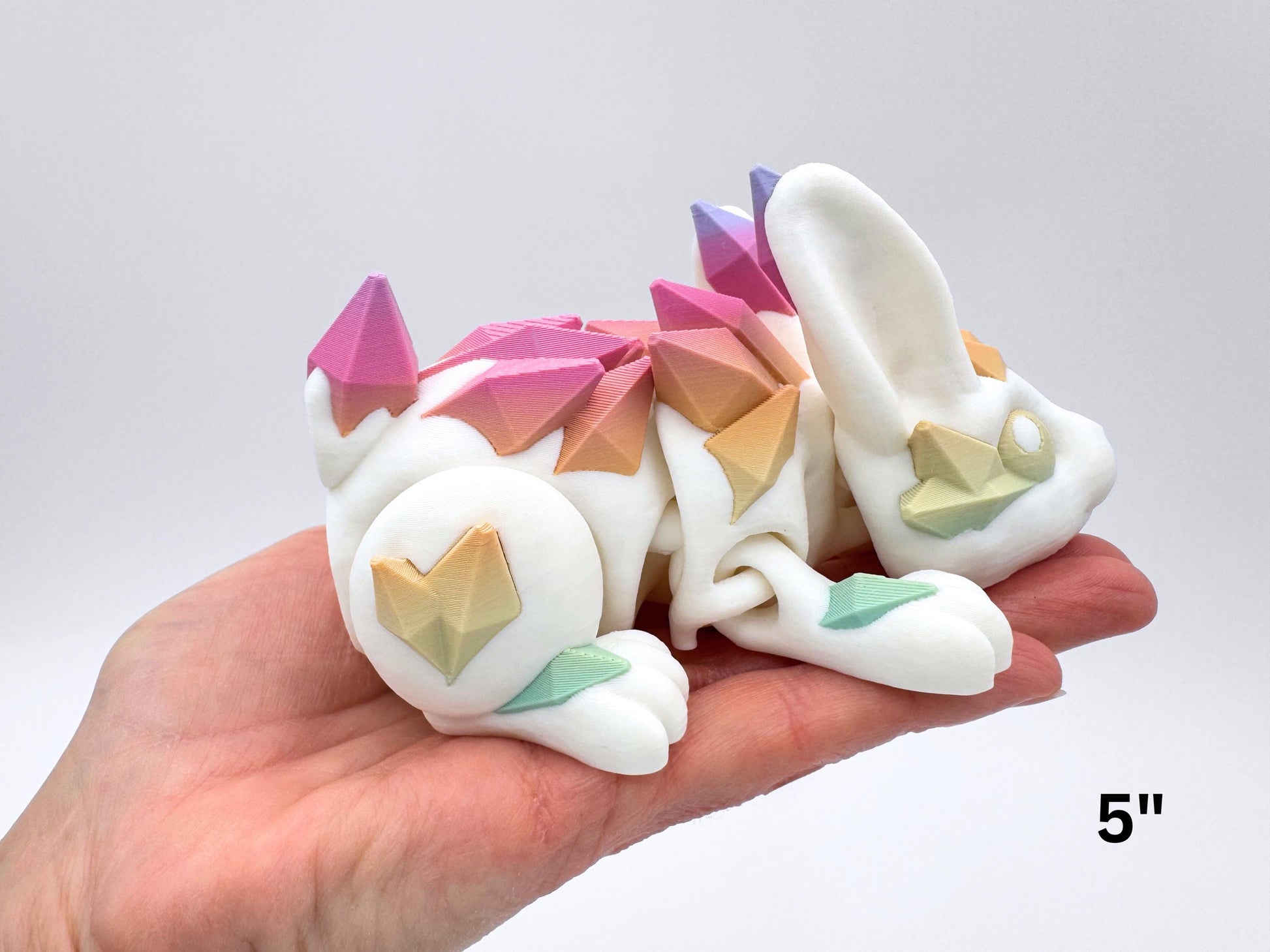 Medium 5" articulated Crystal Bunny displayed on an adult hand for scale. The 3D printed white figure lies on its side with visible hinge joints; faceted crystal plates in soft rainbow colors run along the back, ear, cheek, forearm, and a heart-shaped plate on the thigh. Lighting is even on a neutral gray background. Overlay text at lower right reads “5".