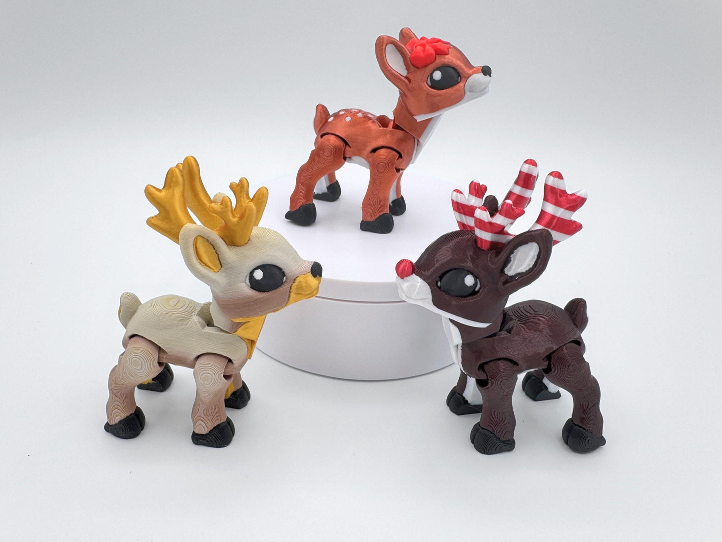 Three 3D printed articulated reindeer figurines in different styles are displayed together, each with flexible joints. One reindeer is tan with golden antlers, another is dark brown with red and white striped candy cane antlers, and the third is copper red with a red bow. These small reindeer figurines work as Christmas collectibles, holiday decorations, or playful articulated fidgets.