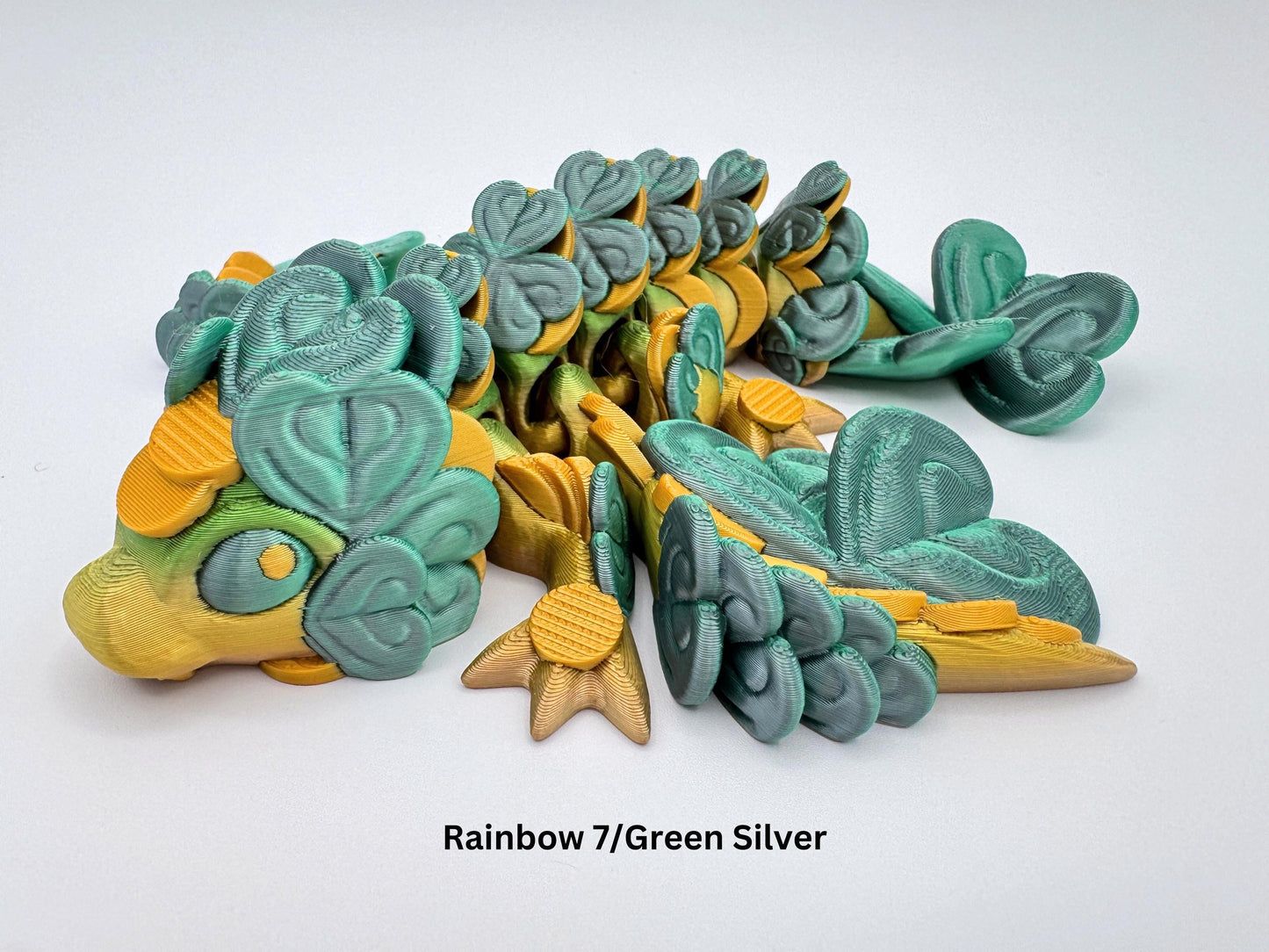 A close-up side view of a baby clover dragon figurine with a rounded snout and large, clover-shaped leaf details covering the head, legs, back and tail. The dragon's eye is visible and surrounded by small raised shapes. Its front legs are bent, and the layers of clover leaves create a textured, leafy appearance. The angle shows the overlapping scale like patterns from head to tail in detail.