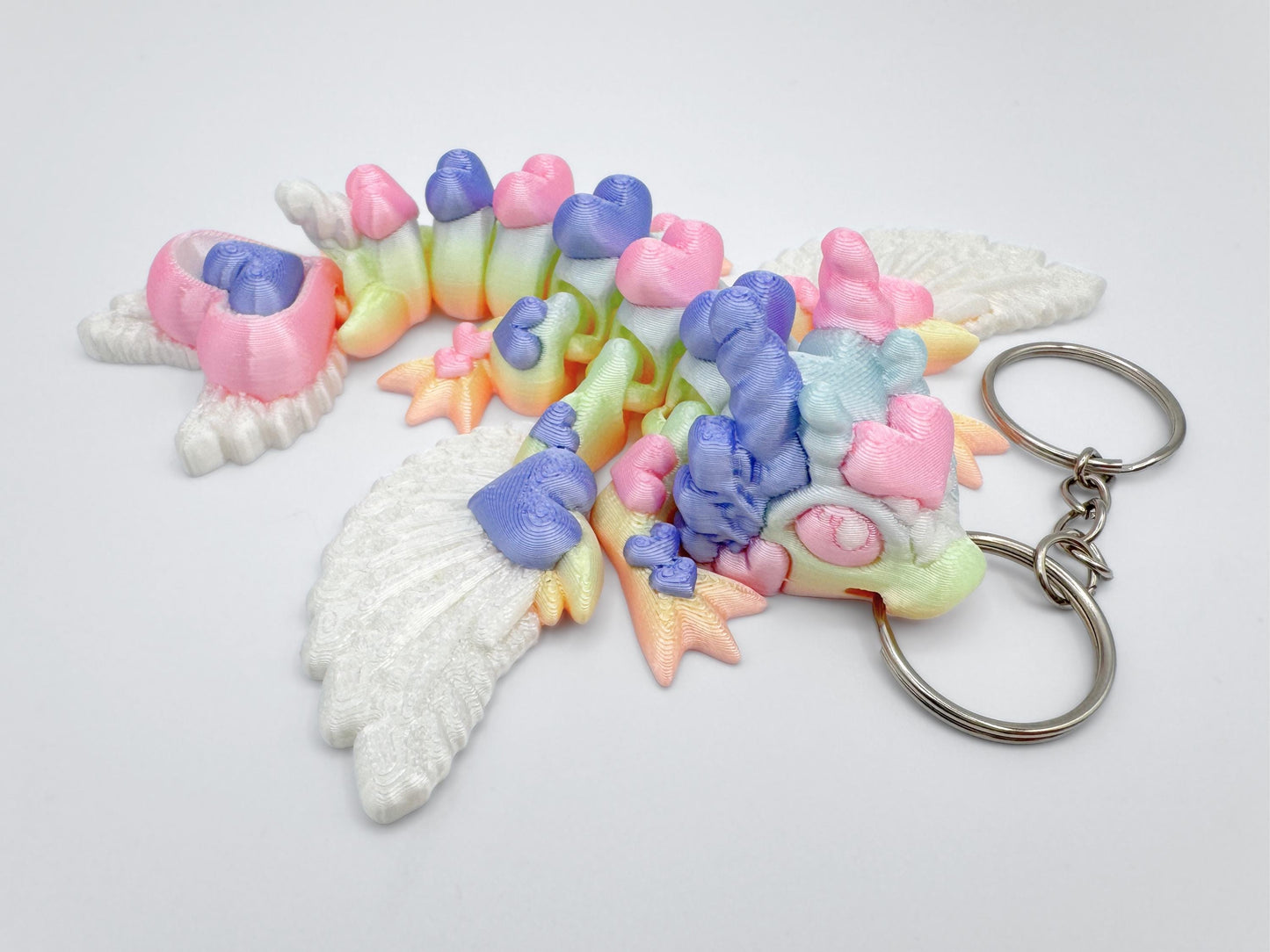 Top-down view of an articulated  3d printed heart dragon keychain lying flat on a white surface. The dragon’s body is extended in a slight curve, showing heart-shaped joints, a heart-stacked tail, and four small legs. Its head has a rounded snout and ear-like details. The wings are white with textured feather patterns. The body displays visible pastel color transitions, including pink, lavender, blue, mint, and yellow. A silver keyring is attached at the mouth.