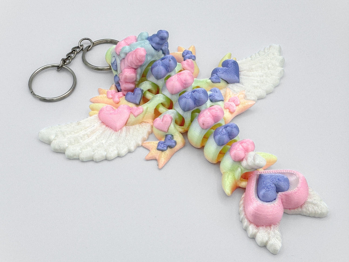 Side view of an articulated 3D printed heart dragon keychain resting on a flat white background. The dragon has white feather-style wings, four legs with small rounded feet, and a tail ending in two heart shapes. Its body is composed of curved heart-shaped links showing soft pastel shades including pink, lavender, mint green, and yellow. The head has ear-like side details and a rounded nose. A silver keyring is attached through the dragon’s snout.