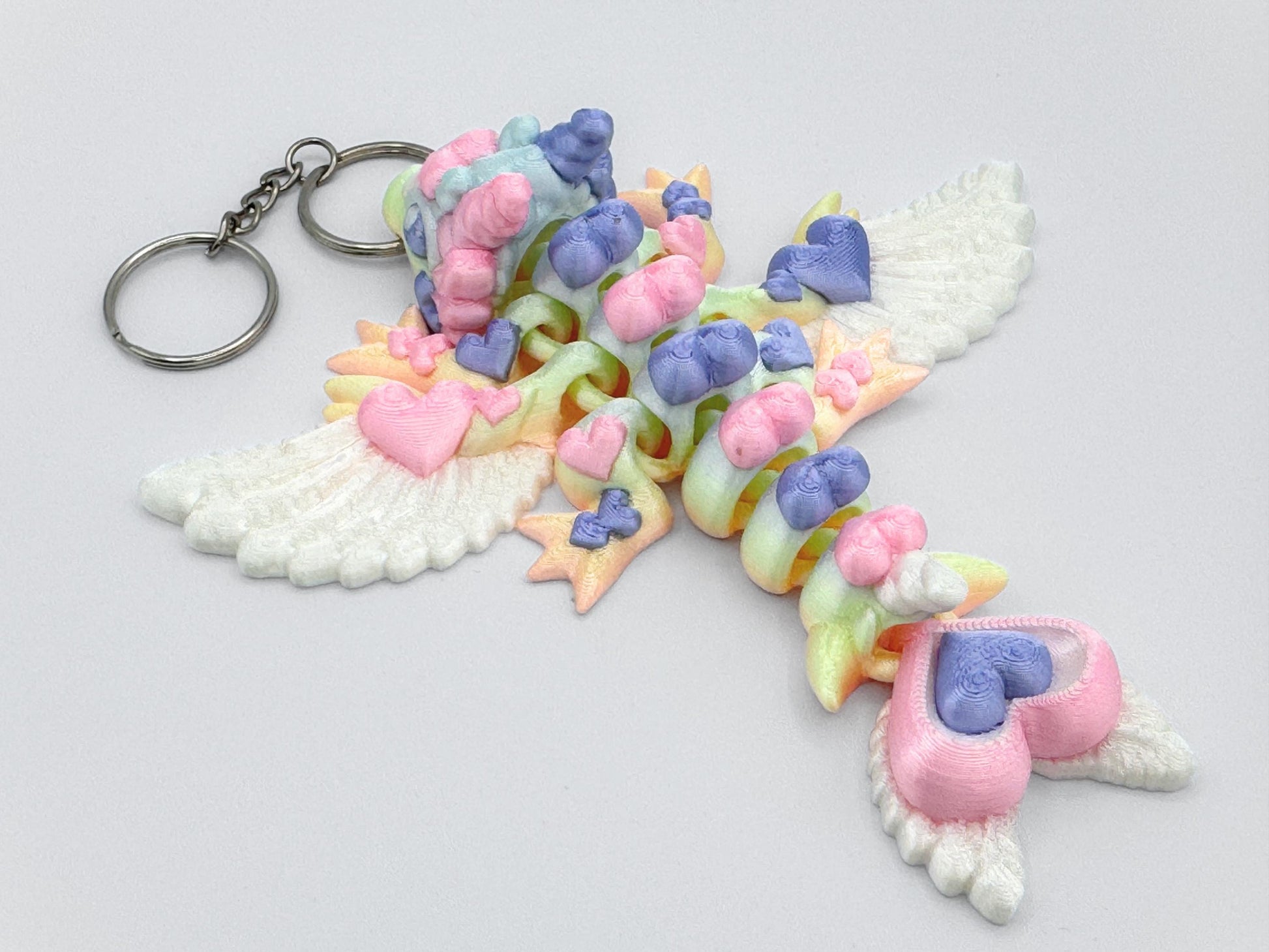 Side view of an articulated 3D printed heart dragon keychain resting on a flat white background. The dragon has white feather-style wings, four legs with small rounded feet, and a tail ending in two heart shapes. Its body is composed of curved heart-shaped links showing soft pastel shades including pink, lavender, mint green, and yellow. The head has ear-like side details and a rounded nose. A silver keyring is attached through the dragon’s snout.
