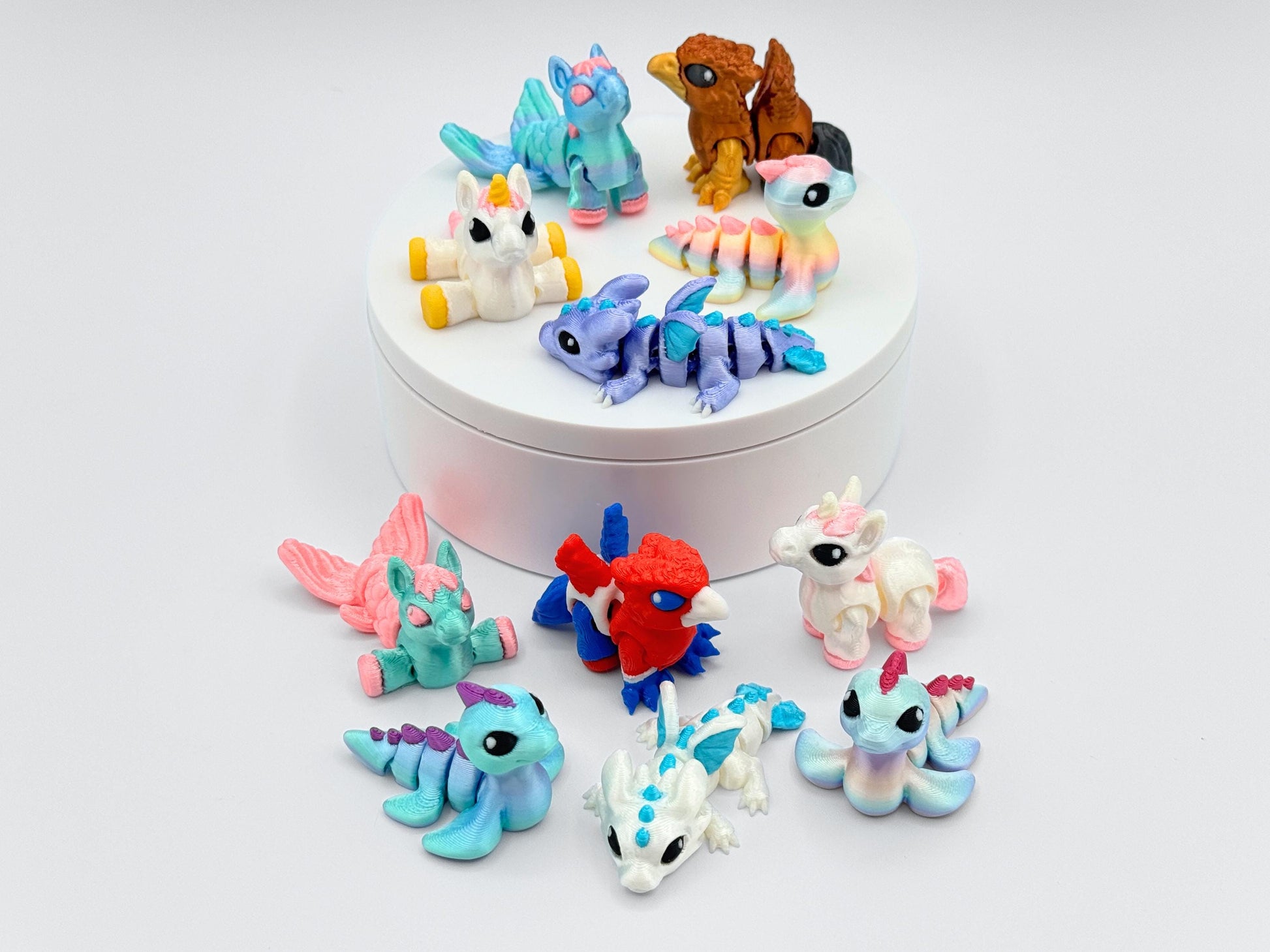 Tiny Creatures Animal Figurines 3D printed arranged around a white round stand, including unicorns, dragons, gryphons, and sea-inspired figures in pastel, rainbow, and metallic colors. Each figurine is detailed with smooth articulated segments, vibrant accents, and expressive black eyes, shown from above on a clean white background.