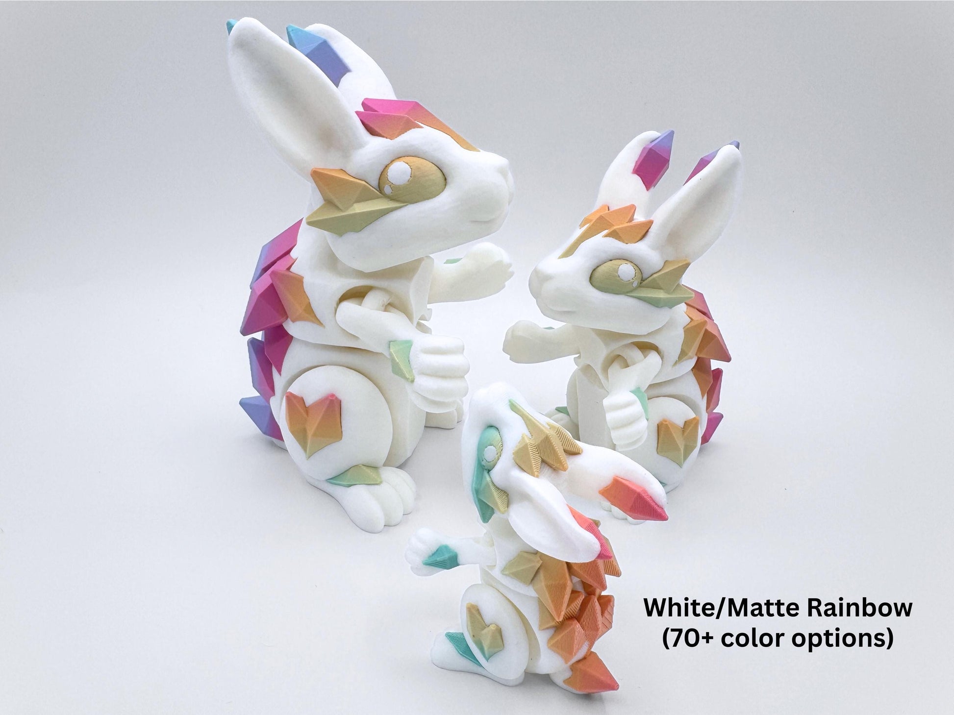 3D printed articulated crystal bunnies with white bodies, vibrant pastel rainbow crystal spikes, and crystal-shaped accents in pink, purple, teal, yellow and blue. Features intricate detailing, expressive poses, and smooth, flexible designs. Each white bunny has faceted crystal plates in matte pastel rainbow shades along the ears, cheeks, back, legs, and tail. One large, one medium, and one small bunny face each other; overlay text at lower right reads “White/Matte Rainbow (70+ color options).