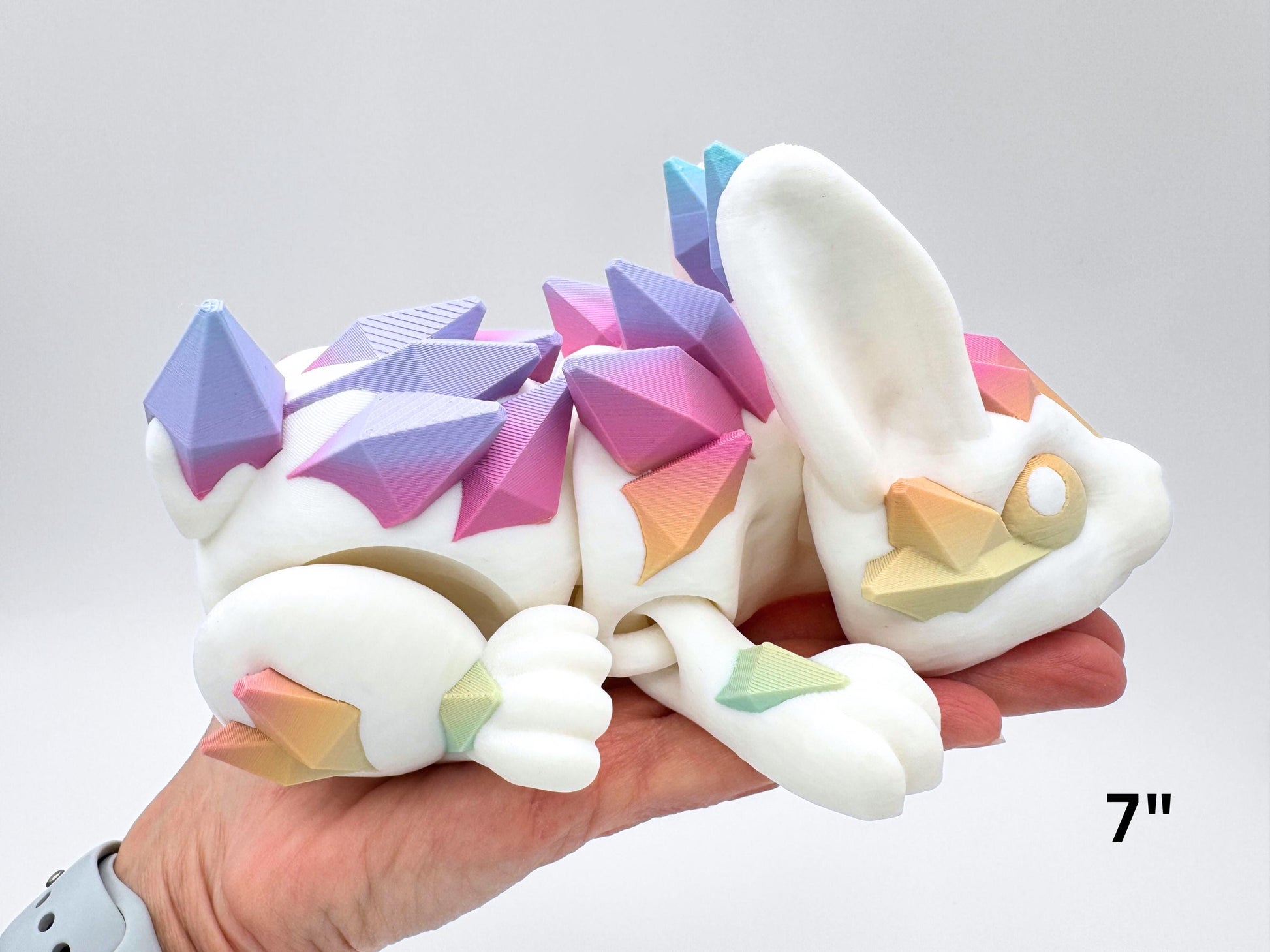 Close view of the Large 7" articulated Crystal Bunny resting across an adult hand for scale. The bunny is 3D printed in white with matte pastel rainbow crystal plates on the back and ear tip; head turned slightly, paws tucked, body in a reclining pose showing segmented joints and layered print texture. Overlay text in the lower right corner reads “7".