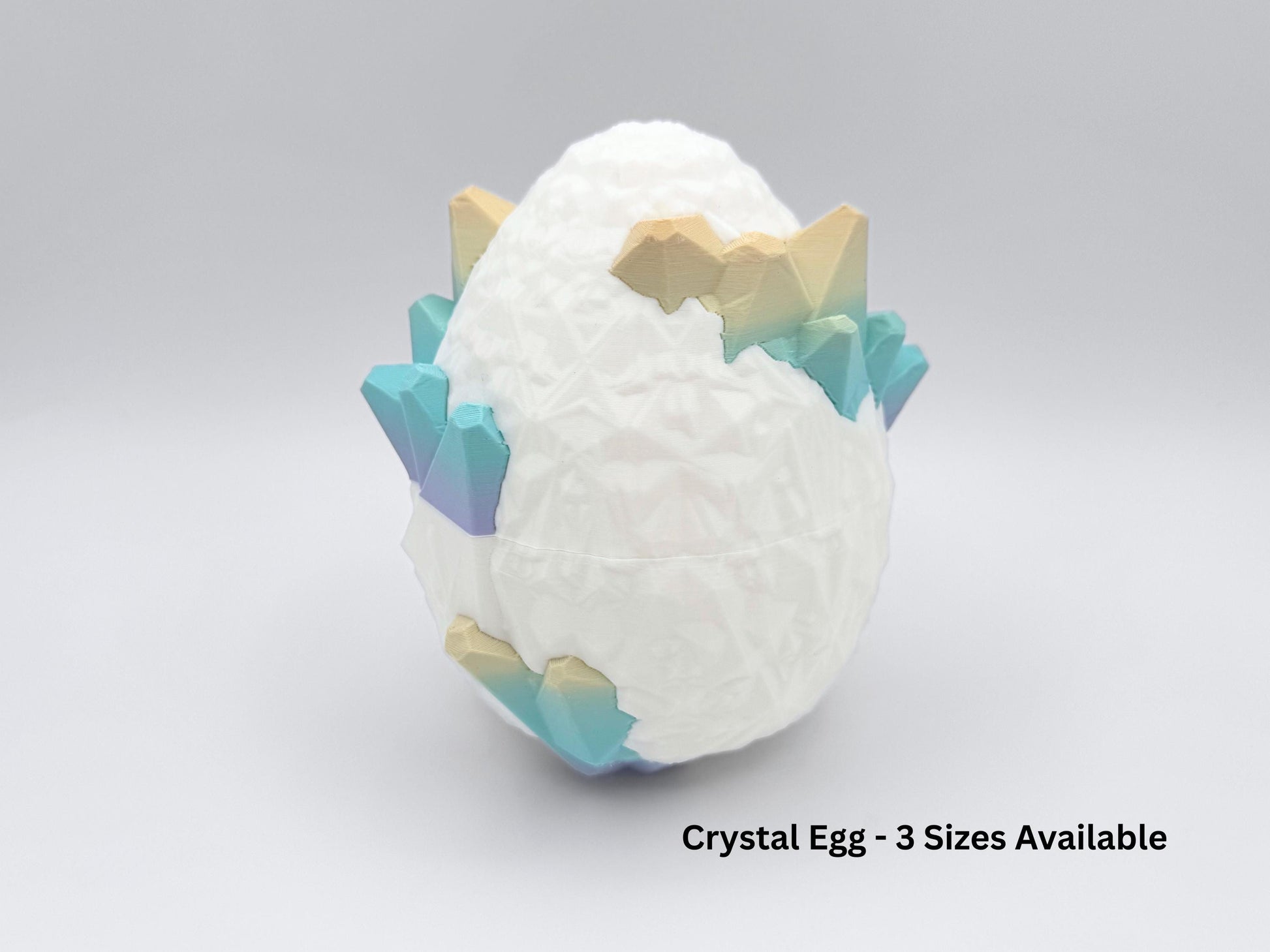 Crystal Egg container shown closed, 3D printed in white with a faceted surface and matte pastel rainbow crystal clusters embedded around the shell, including teal, gold, and lavender gradients. The seam of the two halves is visible around the middle. Neutral studio background. Overlay caption at lower right states “Crystal Egg – 3 Sizes Available.”