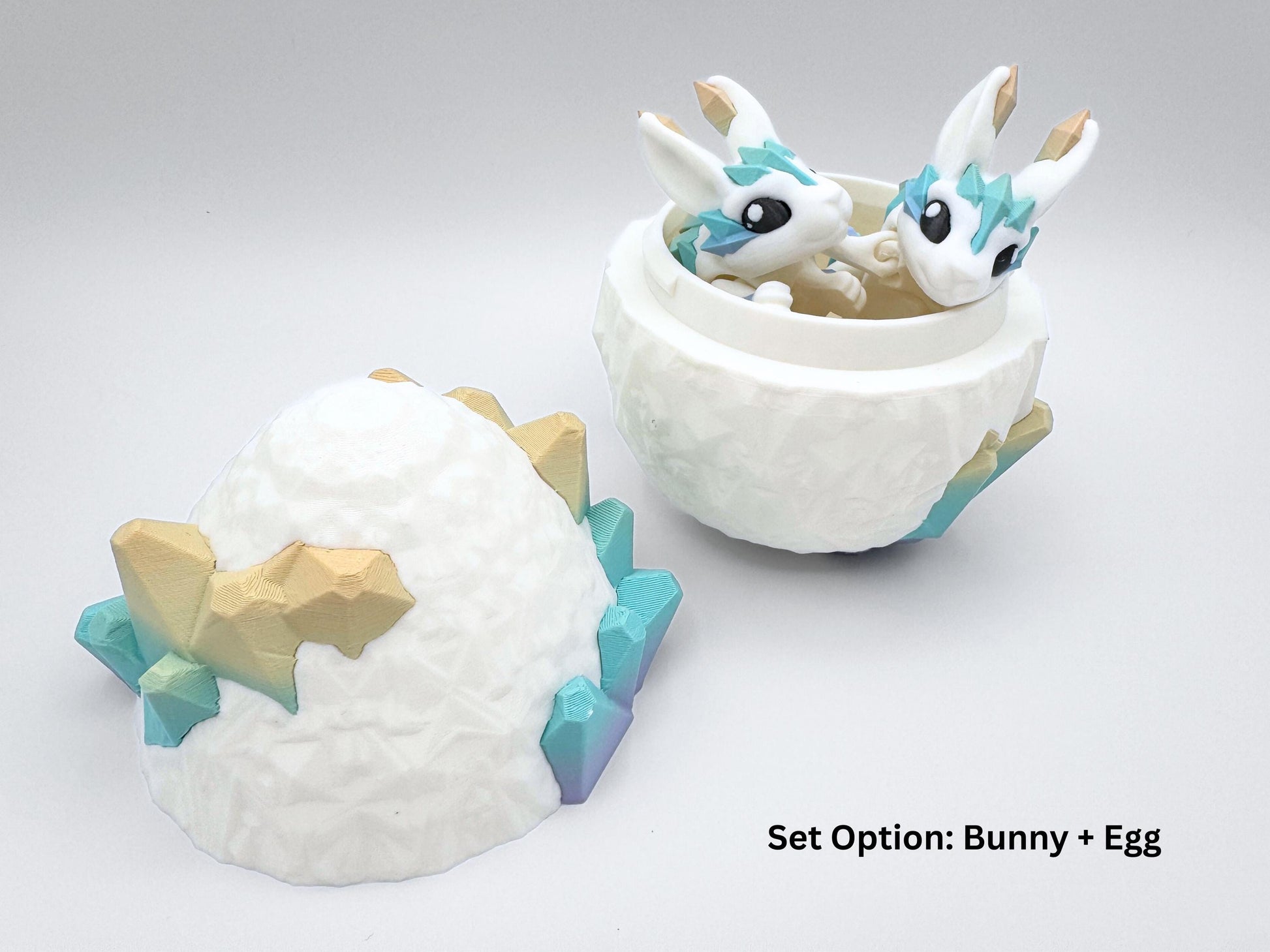 Open Crystal Egg container with two matching mini Crystal Bunny figures nested inside. All pieces are 3D printed; the egg shows a faceted white interior and exterior with pastel rainbow crystal clusters. The articulated bunnies are white with teal-to-gold crystal accents and pointed ear tips. Left half of the egg lies on its side. Overlay caption reads “Set Option: Bunny + Egg.”