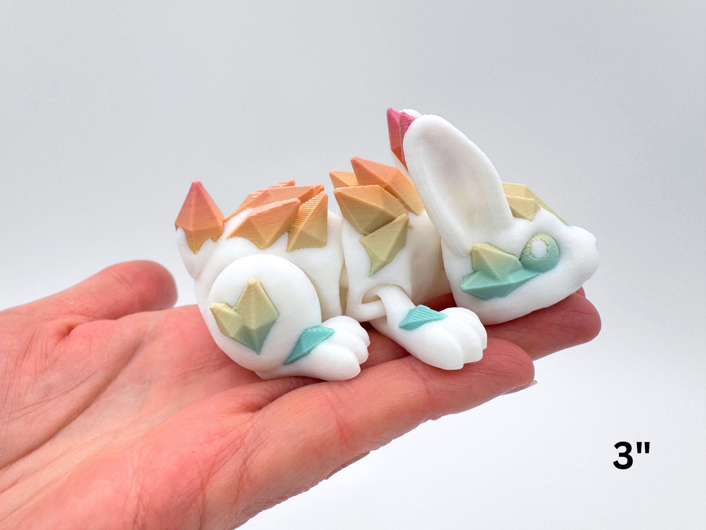 Small 3" articulated Crystal Bunny shown in a palm for size reference. The 3D printed white bunny reclines with compact proportions; pastel rainbow crystal plates accent the ear tip, cheek, foreleg, back, thigh, and tail. Layer lines and segment breaks indicate articulation. Background is smooth light gray. Overlay text in the corner reads “3".