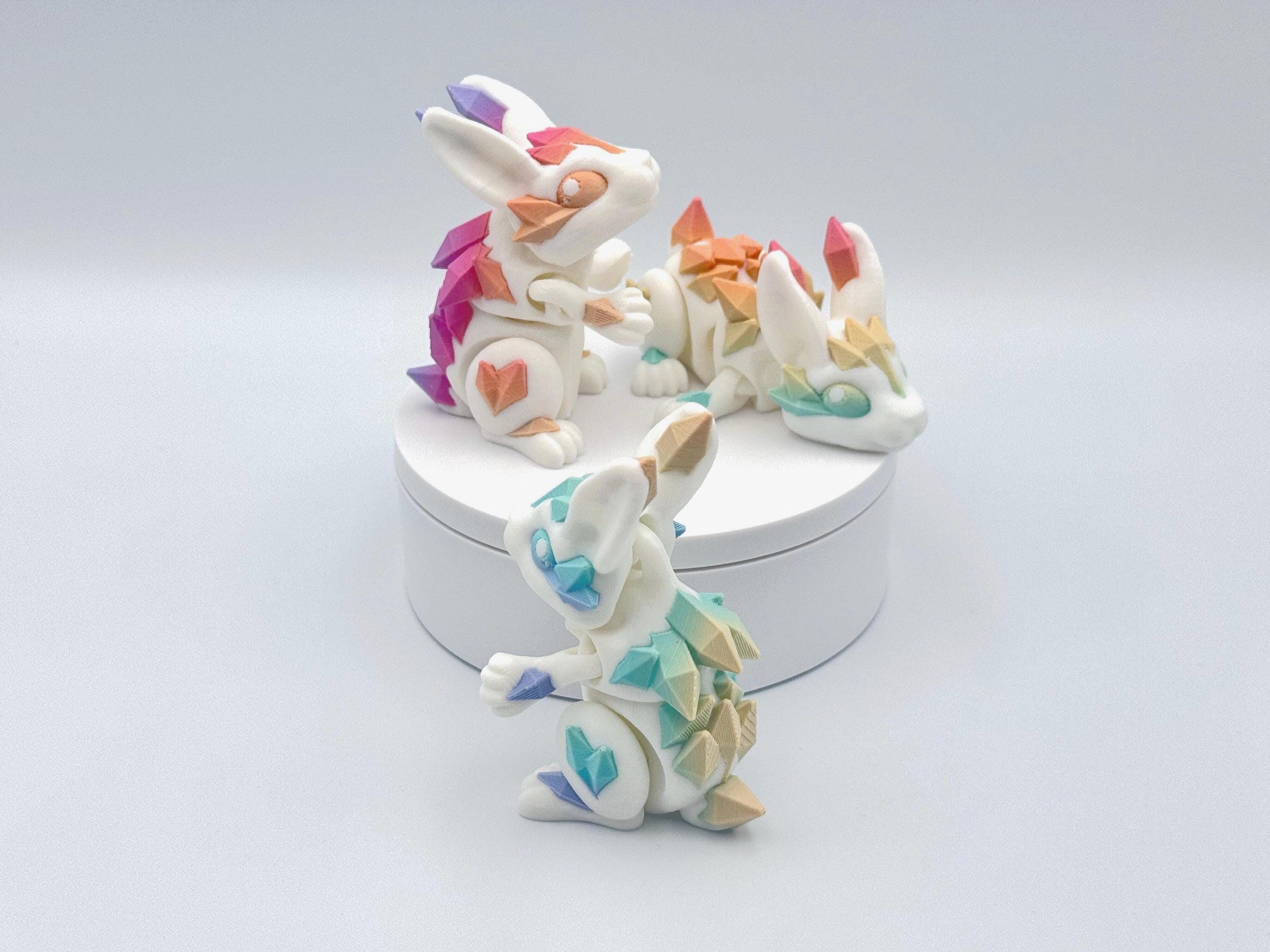 3D printed articulated crystal bunnies with white bodies, vibrant pastel rainbow crystal spikes, and crystal-shaped accents in pink, purple, teal, yellow and blue. Features intricate detailing, expressive poses, and smooth, flexible designs.