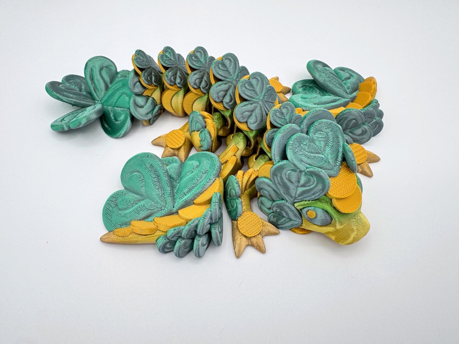 3D printed Baby Clover Dragon lying curled on a flat white surface. The dragon is covered entirely in overlapping clover-shaped leaves, including the head, back, wings and tail. Its limbs are tucked under its body, and the tail curves around in a relaxed pose. The overall shape is compact, with layered textures and rounded forms clearly visible across the figure.