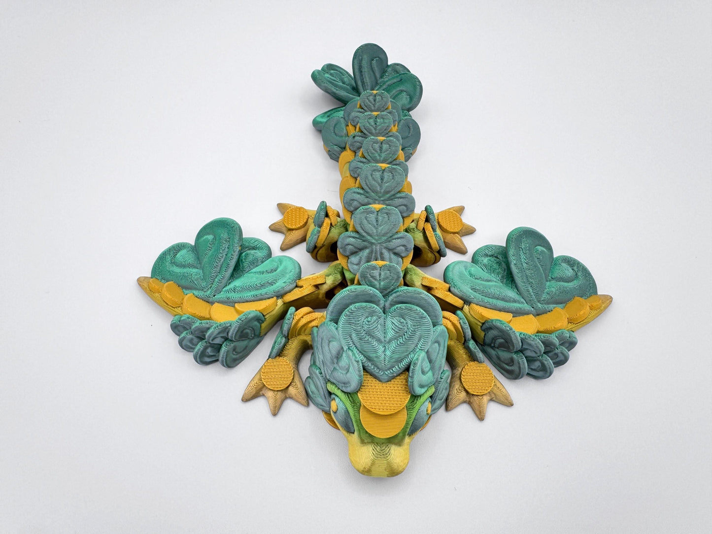 3D printed baby clover dragon figurine displayed flat on a white background, viewed directly from above. The dragon’s limbs are stretched outward, and the full body is covered with clover leaf shapes, including the wings, spine, tail, head and feet. The symmetrical layout shows how the clover leaves form the dragon’s structure in repeating patterns. The head is at the bottom center and the tail tapers toward the top of the image.