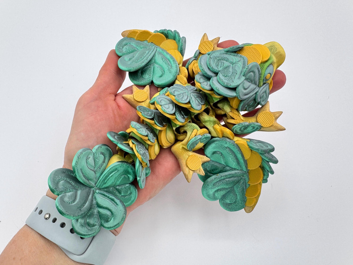 A hand holding a baby clover dragon figurine resting across the palm. The dragon’s body is covered with large, clover-shaped leaves from head to tail. Its legs, wings and tail curve outward, and the full length of the dragon is visible stretched across the hand, showing its size and flexible structure.