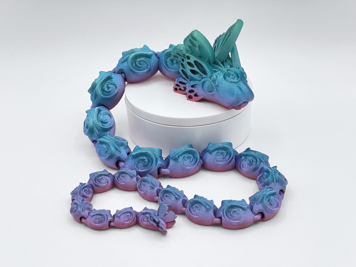 3D printed articulated butterfly snake with rose-shaped body segments, silky gradient blue teal to purple pink colors, posed in a circle on a white round riser with a second coiled section in front; butterfly-style head fins on a clean white background.