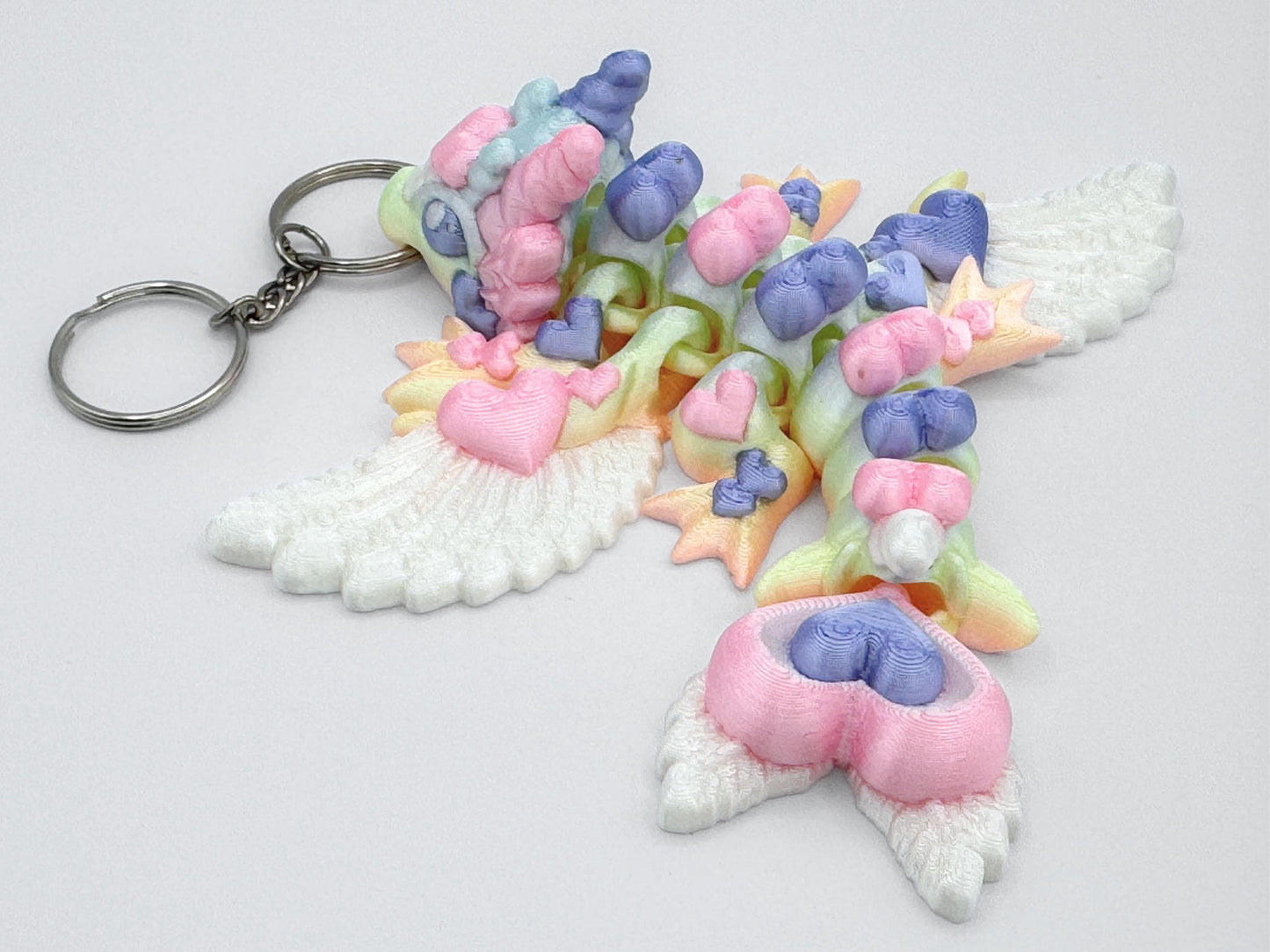 Back-facing view of an articulated 3D printed heart dragon keychain lying flat on a white surface. The dragon’s tail curves to one side and ends in stacked heart shapes. Its back shows a row of heart-shaped body sections in soft colors like pink, purple, yellow, and green. The wings are printed in solid white and have visible feather textures. Four legs and small feet are spread out on both sides. A silver keyring is attached at the head.