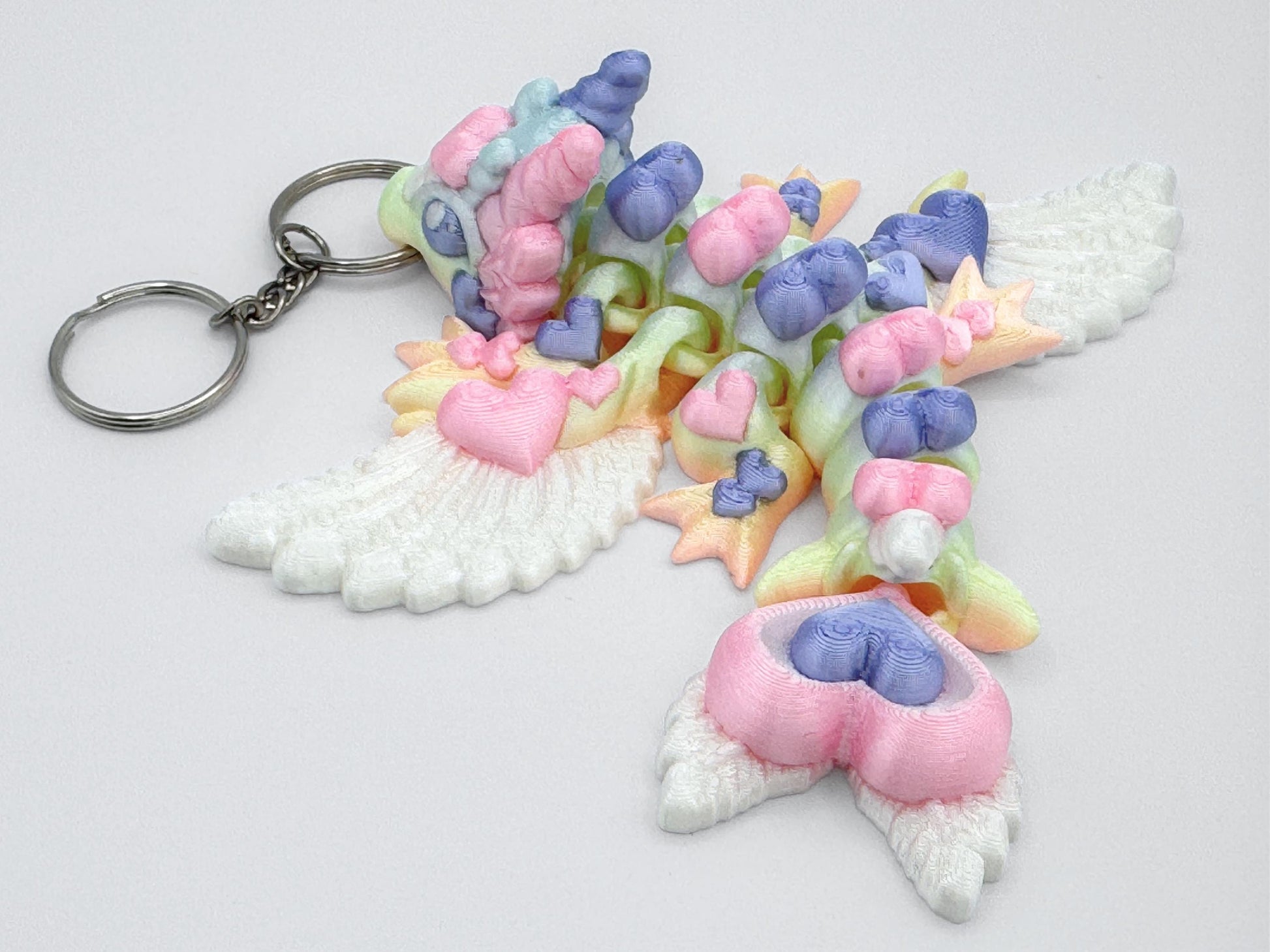 Back-facing view of an articulated 3D printed heart dragon keychain lying flat on a white surface. The dragon’s tail curves to one side and ends in stacked heart shapes. Its back shows a row of heart-shaped body sections in soft colors like pink, purple, yellow, and green. The wings are printed in solid white and have visible feather textures. Four legs and small feet are spread out on both sides. A silver keyring is attached at the head.