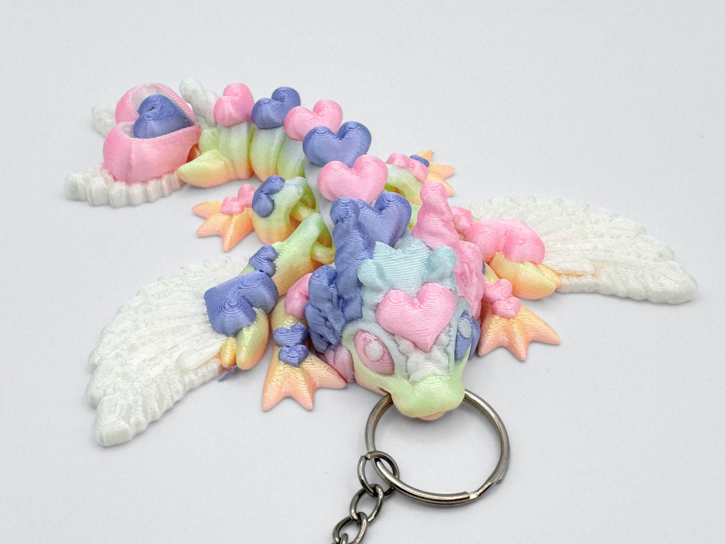 Top-down view of an articulated 3d printed heart dragon keychain lying flat on a white background. The dragon’s body features heart-shaped segments with visible pastel colors, including lavender, pink, mint green, and soft yellow. Its head has two small horn-like ears and a rounded snout. The tail ends in two stacked heart shapes. White feather-textured wings are positioned on the upper body. A silver keyring is inserted at the front of the head.