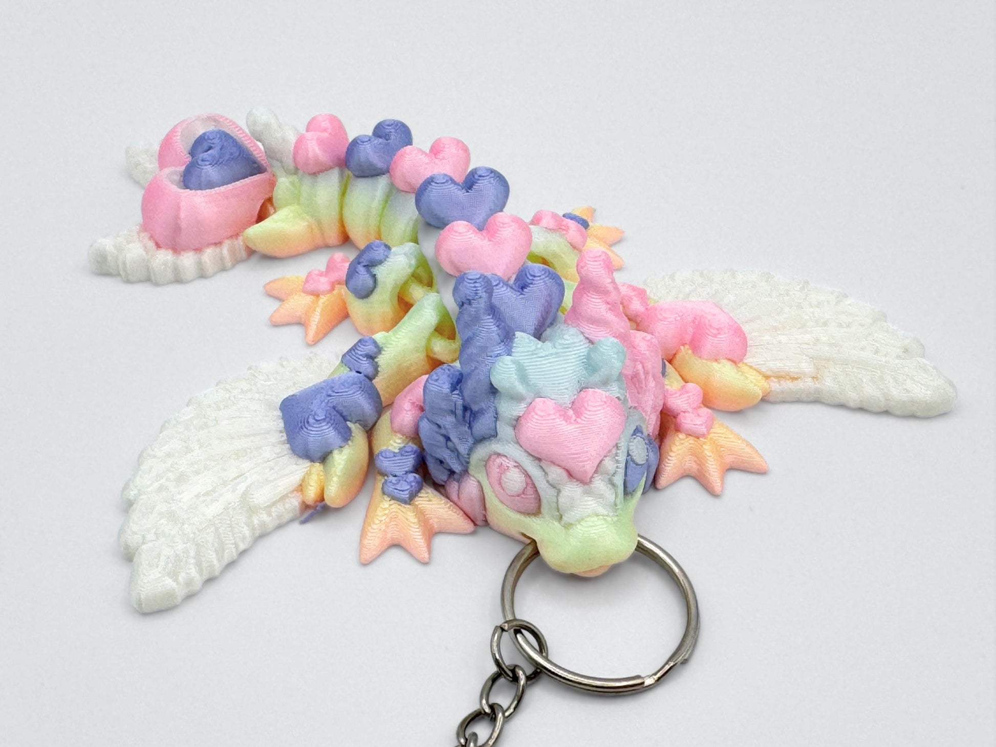 Top-down view of an articulated 3d printed heart dragon keychain lying flat on a white background. The dragon’s body features heart-shaped segments with visible pastel colors, including lavender, pink, mint green, and soft yellow. Its head has two small horn-like ears and a rounded snout. The tail ends in two stacked heart shapes. White feather-textured wings are positioned on the upper body. A silver keyring is inserted at the front of the head.