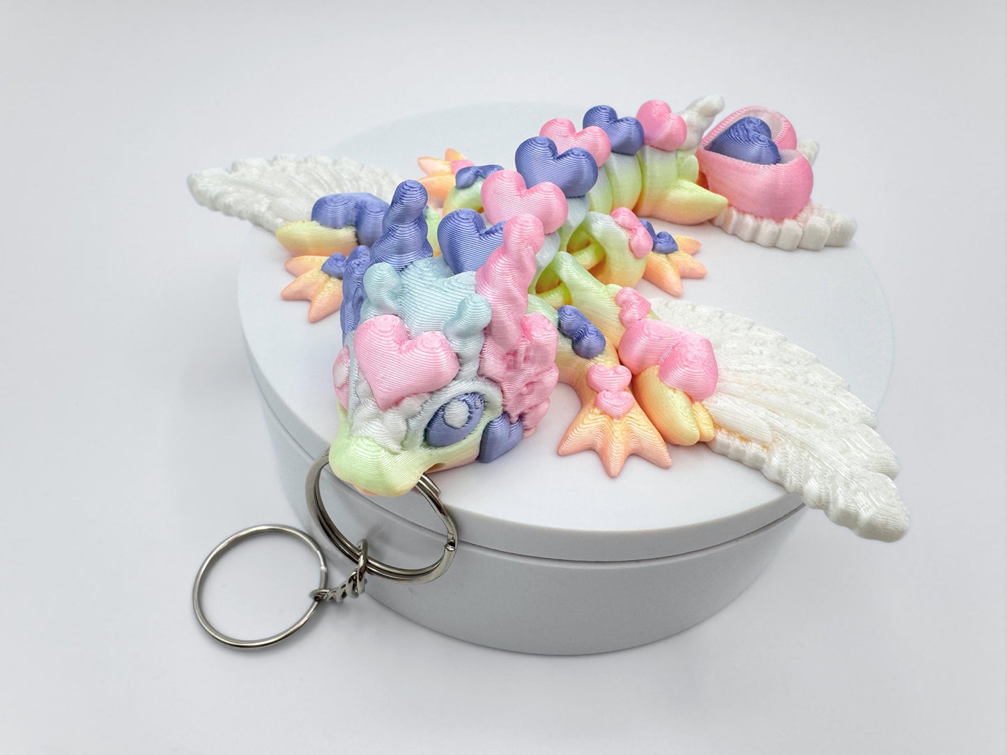 Close-up of an articulated 3D printed heart dragon keychain resting on a round white box. The dragon features white feathered wings, a rounded snout, and short ear-like protrusions. Its long body is made up of heart-shaped segments that curve to the left. The visible colors are pastel pink, mint green, lavender, light blue, and soft yellow arranged in smooth transitions. A silver keyring is attached to the front of the head.
