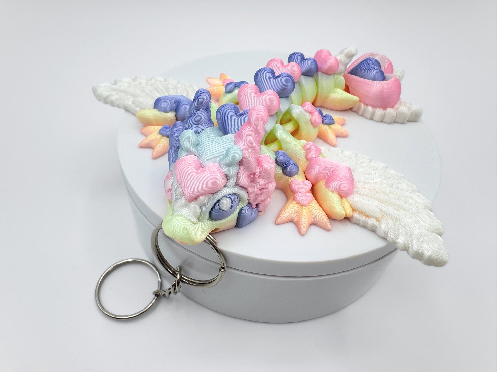 Close-up of an articulated 3D printed heart dragon keychain resting on a round white box. The dragon features white feathered wings, a rounded snout, and short ear-like protrusions. Its long body is made up of heart-shaped segments that curve to the left. The visible colors are pastel pink, mint green, lavender, light blue, and soft yellow arranged in smooth transitions. A silver keyring is attached to the front of the head.