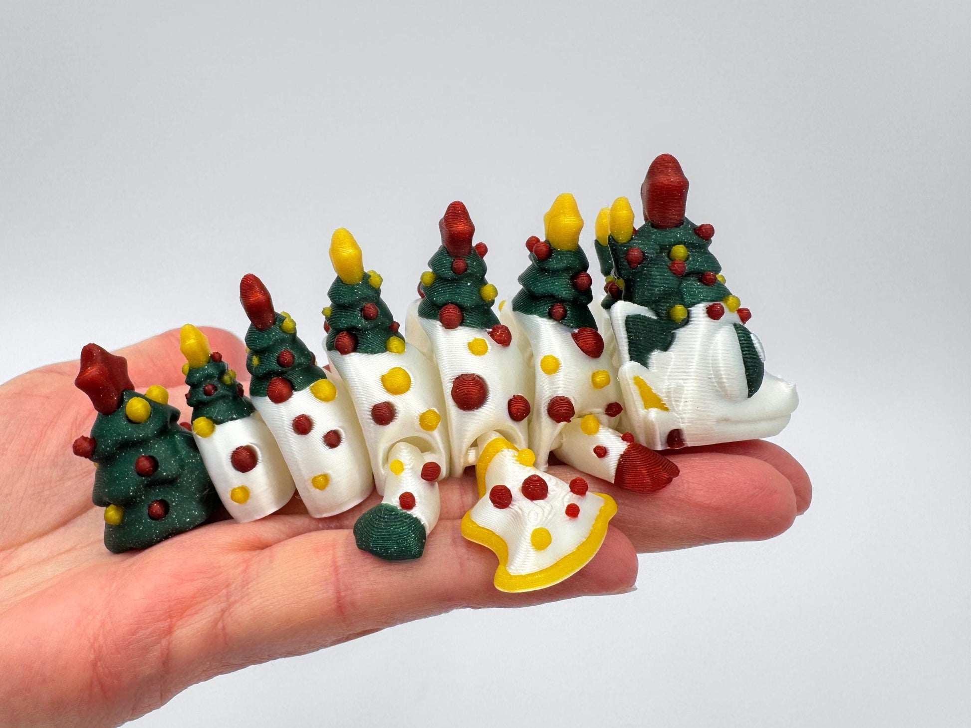 A white 3D printed articulated Christmas tree dragon displayed across a hand, featuring green glitter tree sections with glitter red and yellow ornament details and a red yellow star on each segment. The dragon has articulated linked body parts, painted accents, and holiday-themed colors matching the Christmas dragon and egg tabletop decor set. Neutral white background for clear color reference.