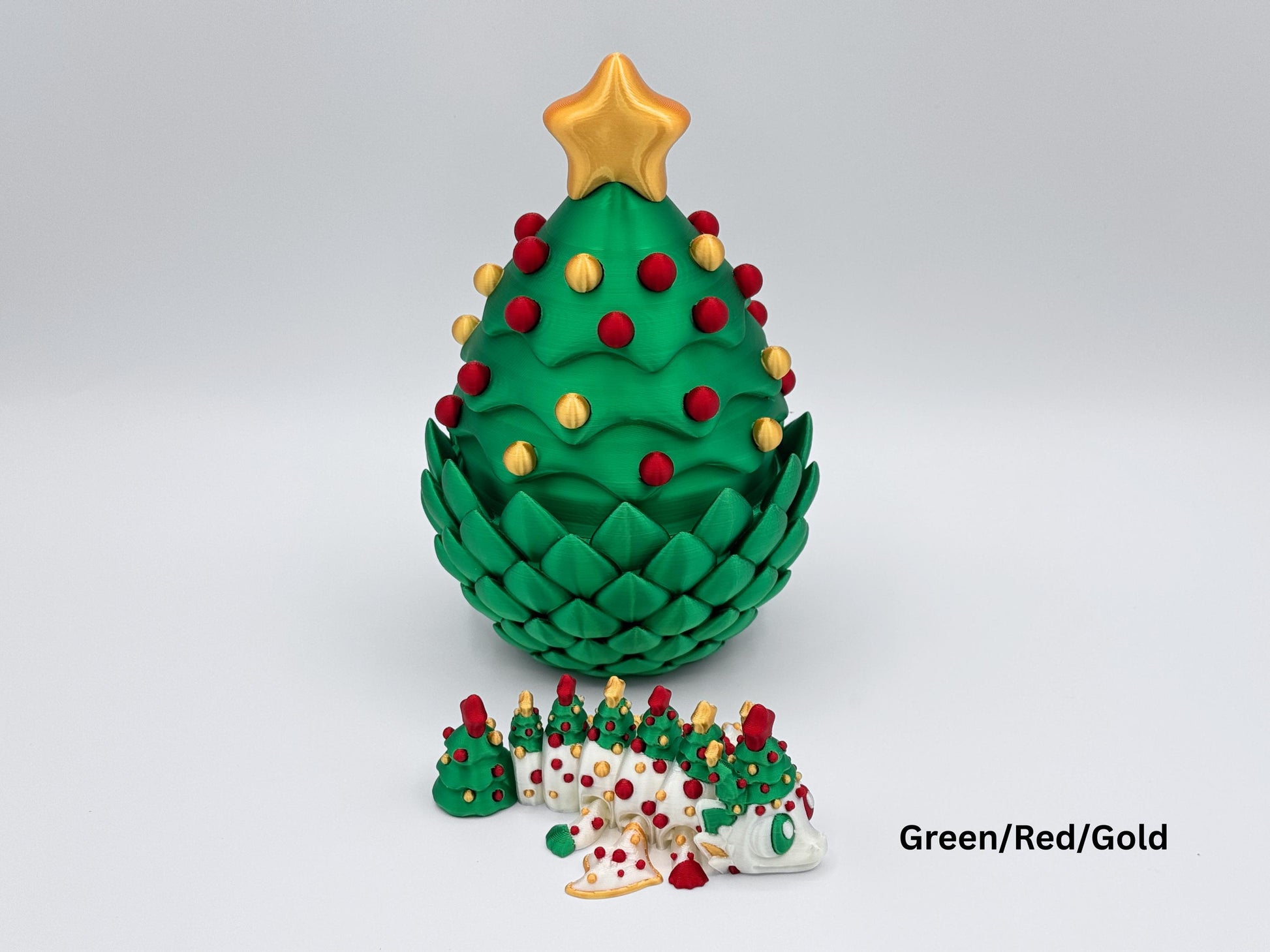 A 3D printed Christmas tree dragon egg in a green, red and gold color, shaped like a layered evergreen with a gold star on top and red-and-gold ornaments, shown with three articulated Christmas dragons in matching colors posed in front on a white background.