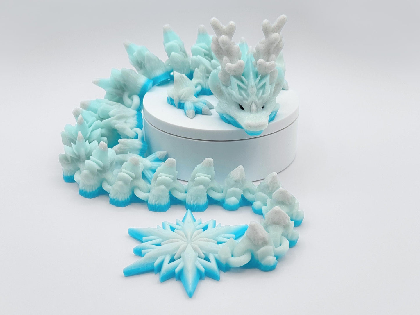3D printed winter dragon figurine with articulated body covered in sharp snow peak textures, large antler horns and icy scales in white glitter. The tail ends in a star shaped snowflake that rests near the box edge as the looped body circles it. The dragon is posed on a white background for full body visibility.
