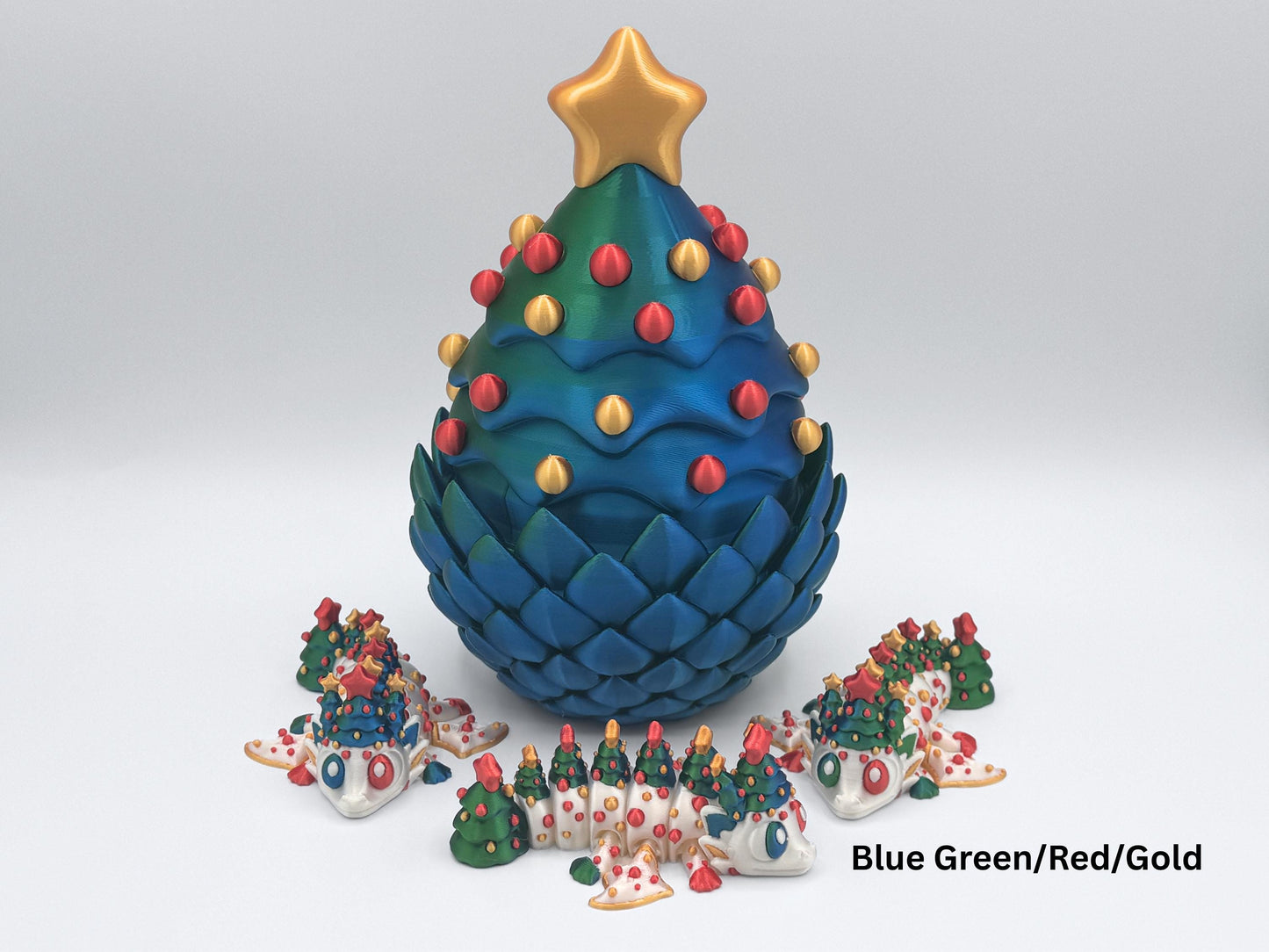 A blue - green 3D printed Christmas tree–shaped dragon egg with gold star topper and red-and-gold ornaments, displayed closed on a white background. In front of the egg are 3 articulated Christmas tree dragons in white filament with blue green trees, red and green ornaments. The image shows the Blue Green/Red/Gold color option for the Christmas dragon and egg set.