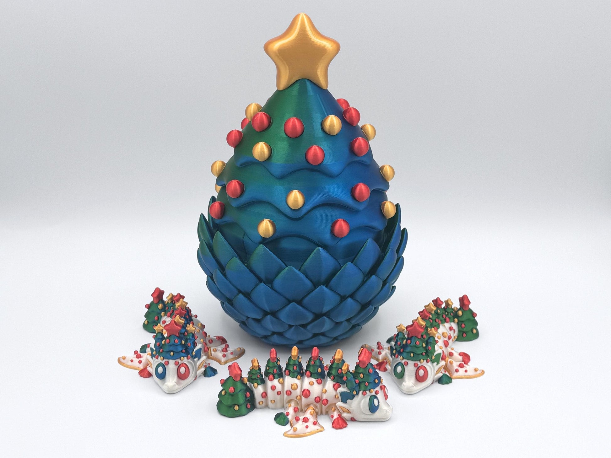 A 3D printed Christmas tree–shaped dragon egg in blue green with a gold star and red-and-gold ornaments, surrounded by three articulated Christmas dragons posed in front. The set is shown on a white background as holiday tabletop decor and a winter centerpiece.